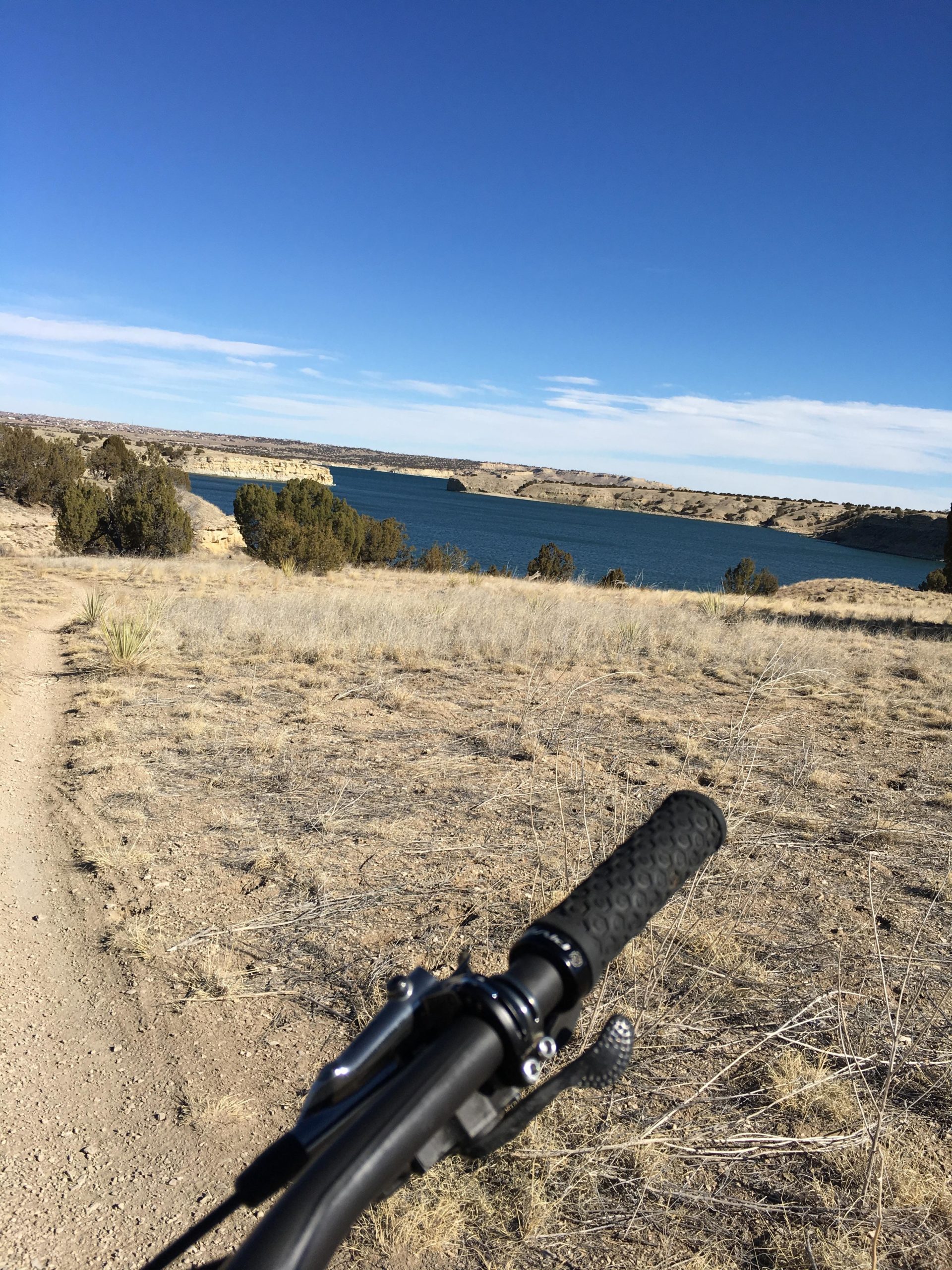 A view from a bike handlebar looking towards a calm blue lake surrounded by rocky hills and sparse vegetation, under a clear blue sky. The dirt path is visible in the foreground, leading toward the water. South Shore Lake Pueblo mountain bike trail.