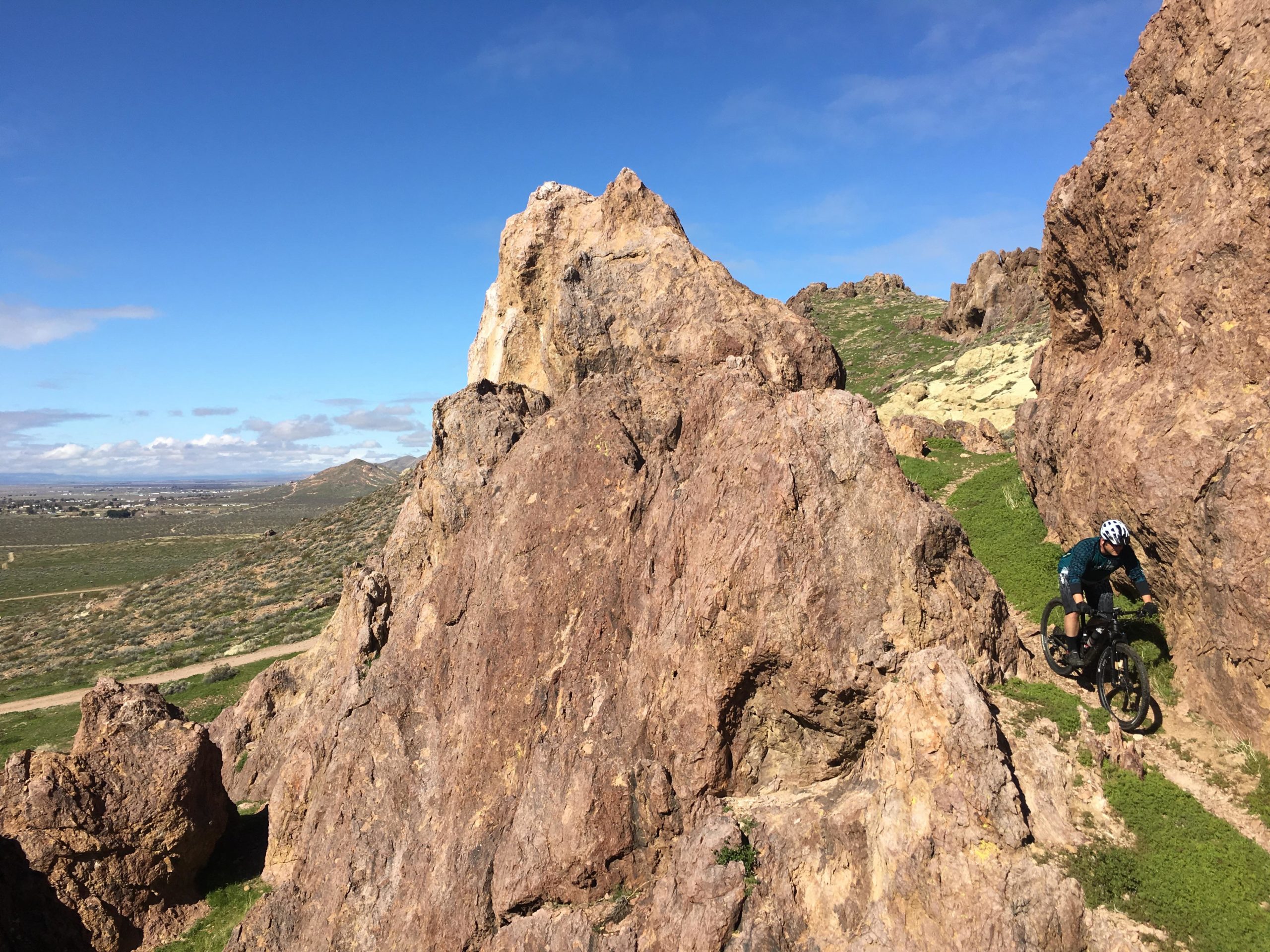 A mountain biker navigates a rocky trail surrounded by rugged terrain and green grass under a clear blue sky. Rosamond Circuit mountain bike trail.