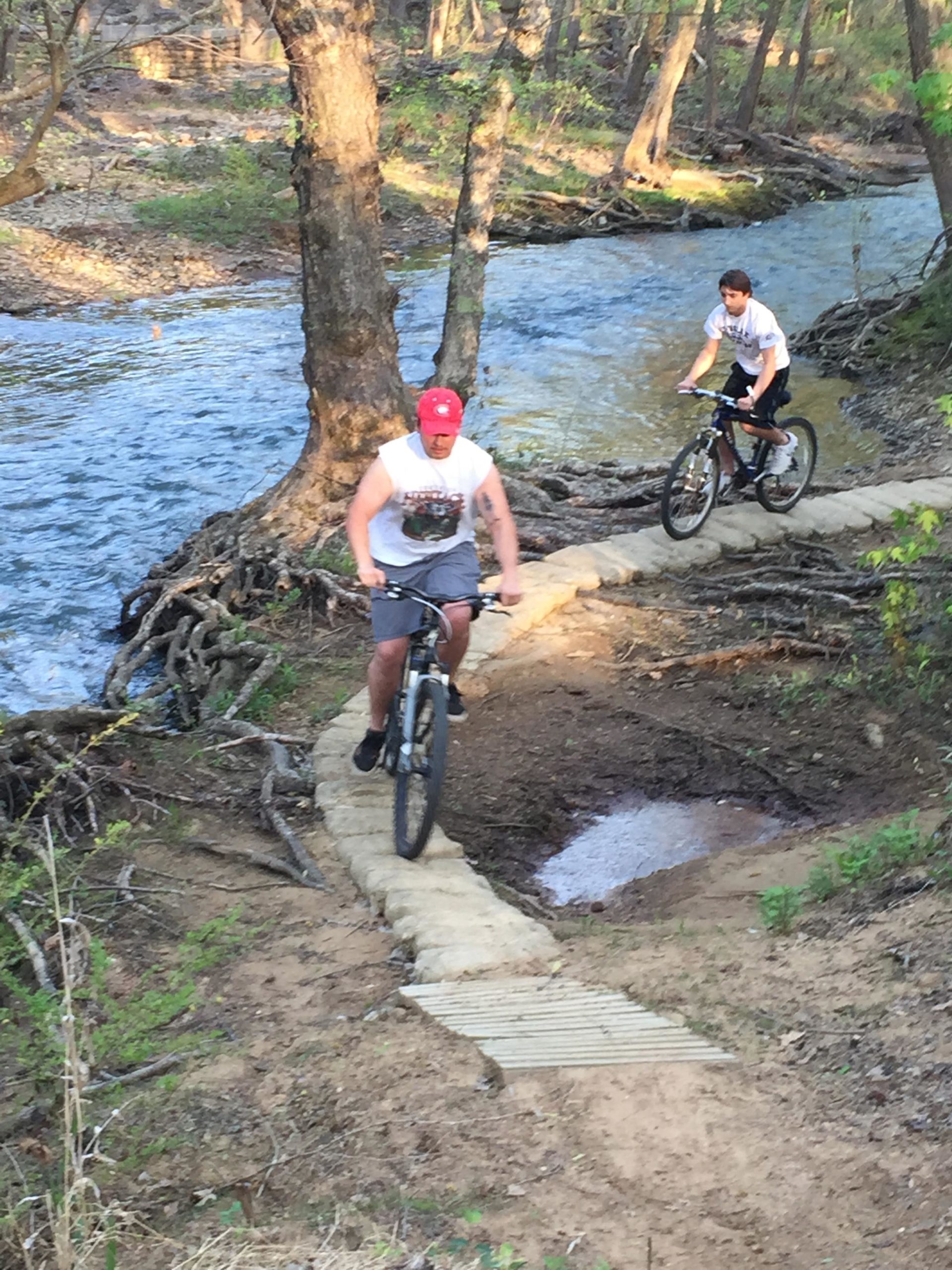 Two individuals are riding bicycles along a narrow dirt path that winds beside a stream in a wooded area. One cyclist, wearing a sleeveless shirt and a red cap, is navigating a section of the trail, while the other, dressed in a t-shirt and shorts, approaches from behind. The surrounding landscape features lush greenery and trees, with the clear water of the stream visible nearby. Spadra Creek Nature Trail mountain bike trail.