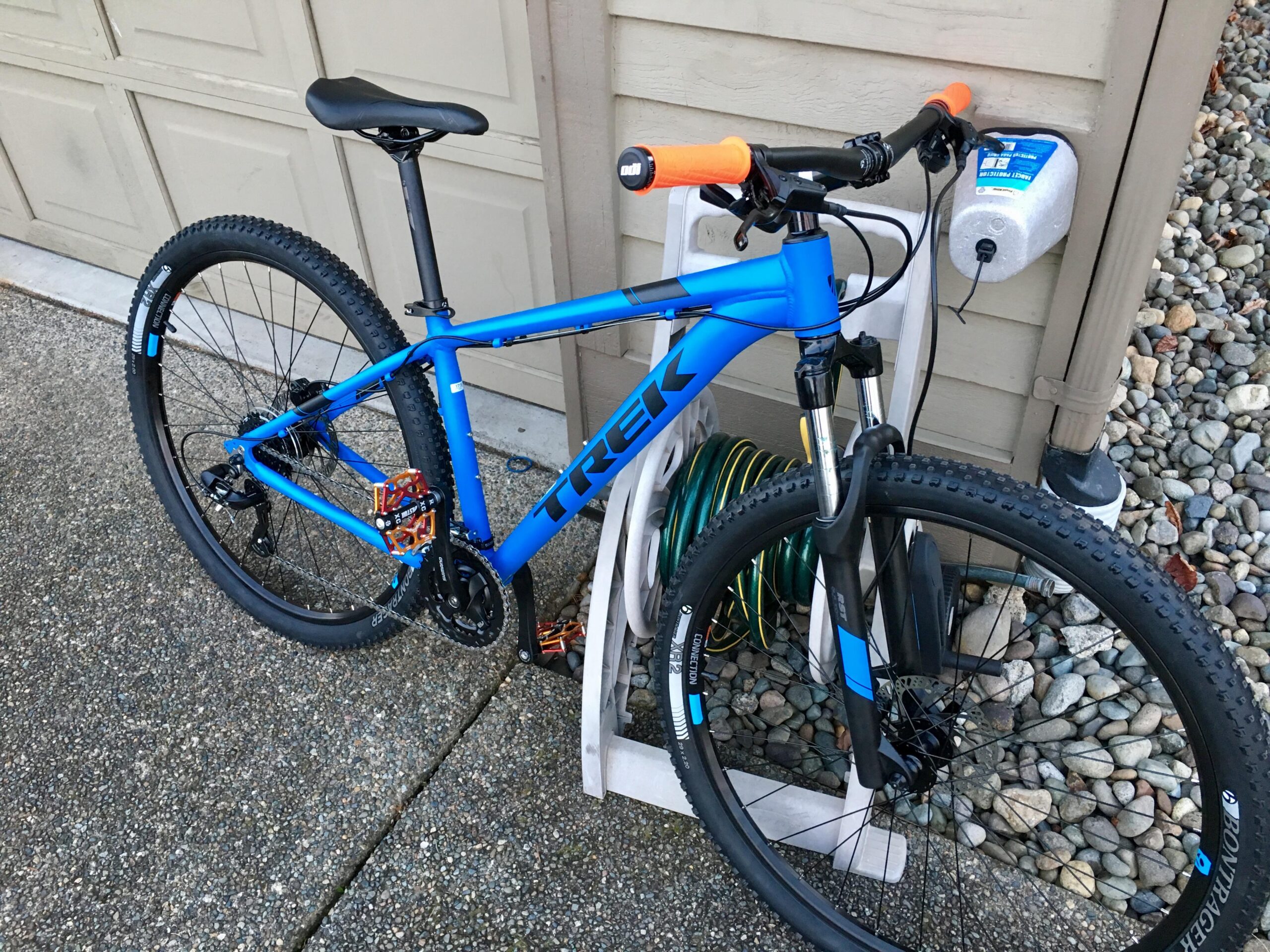 Trek Marin 6: A blue Trek mountain bike positioned next to a garage, featuring orange handlebars and a black seat. The bike has thick, knobby tires and visible gears. In the background, there is a hose reel and a landscaped area with pebbles.