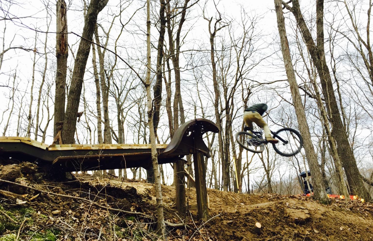 A mountain biker jumping off a wooden ramp in a forested area with bare trees, showcasing action and outdoor sports. North Gorge mountain bike trail.