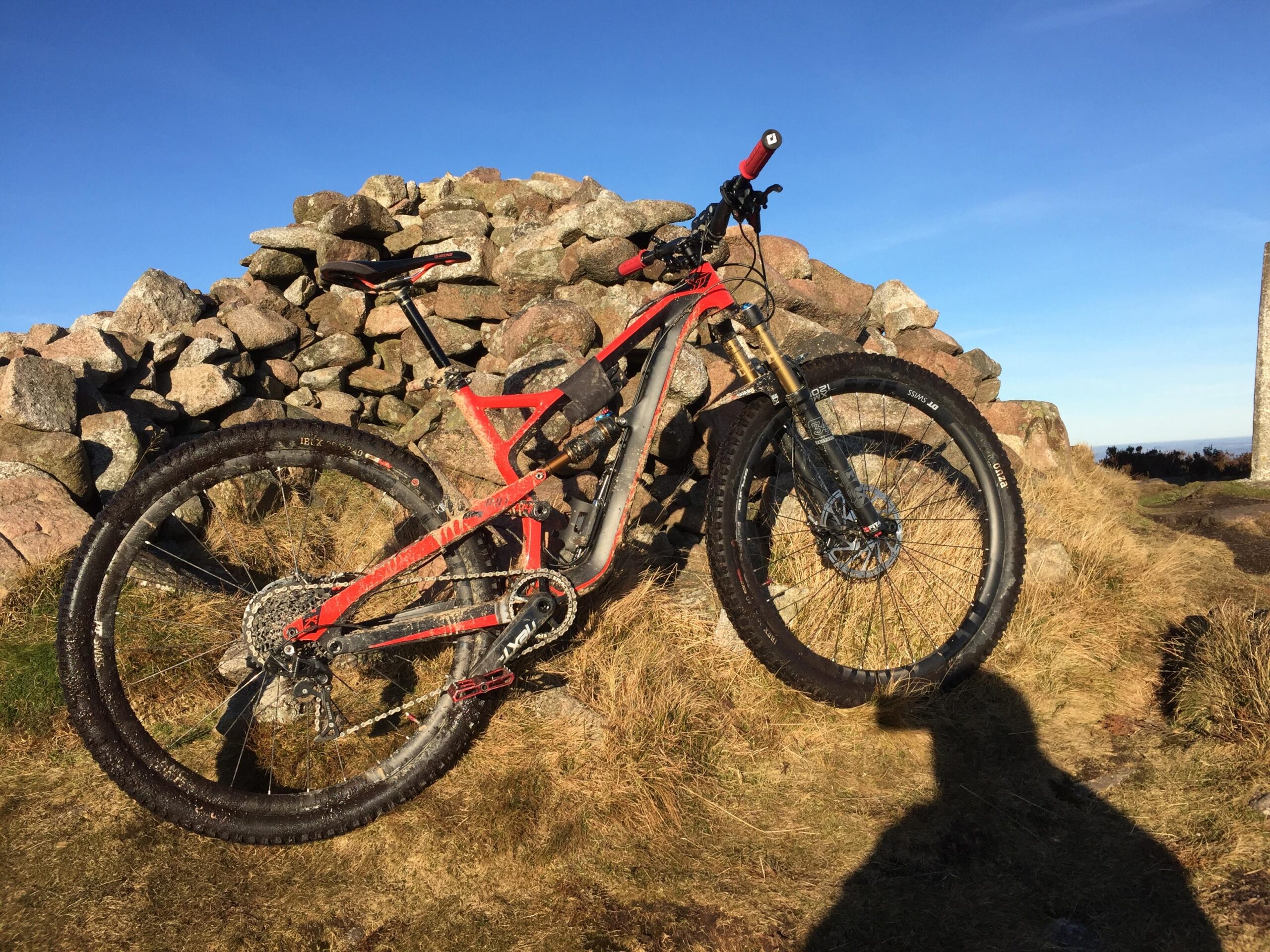 Yt industries Jeffsy: A mountain bike with red accents is leaning against a stone pile on a grassy hill, under a clear blue sky. The bike appears slightly muddy, indicating recent use on rugged terrain. The sun casts a shadow of the bike onto the ground.