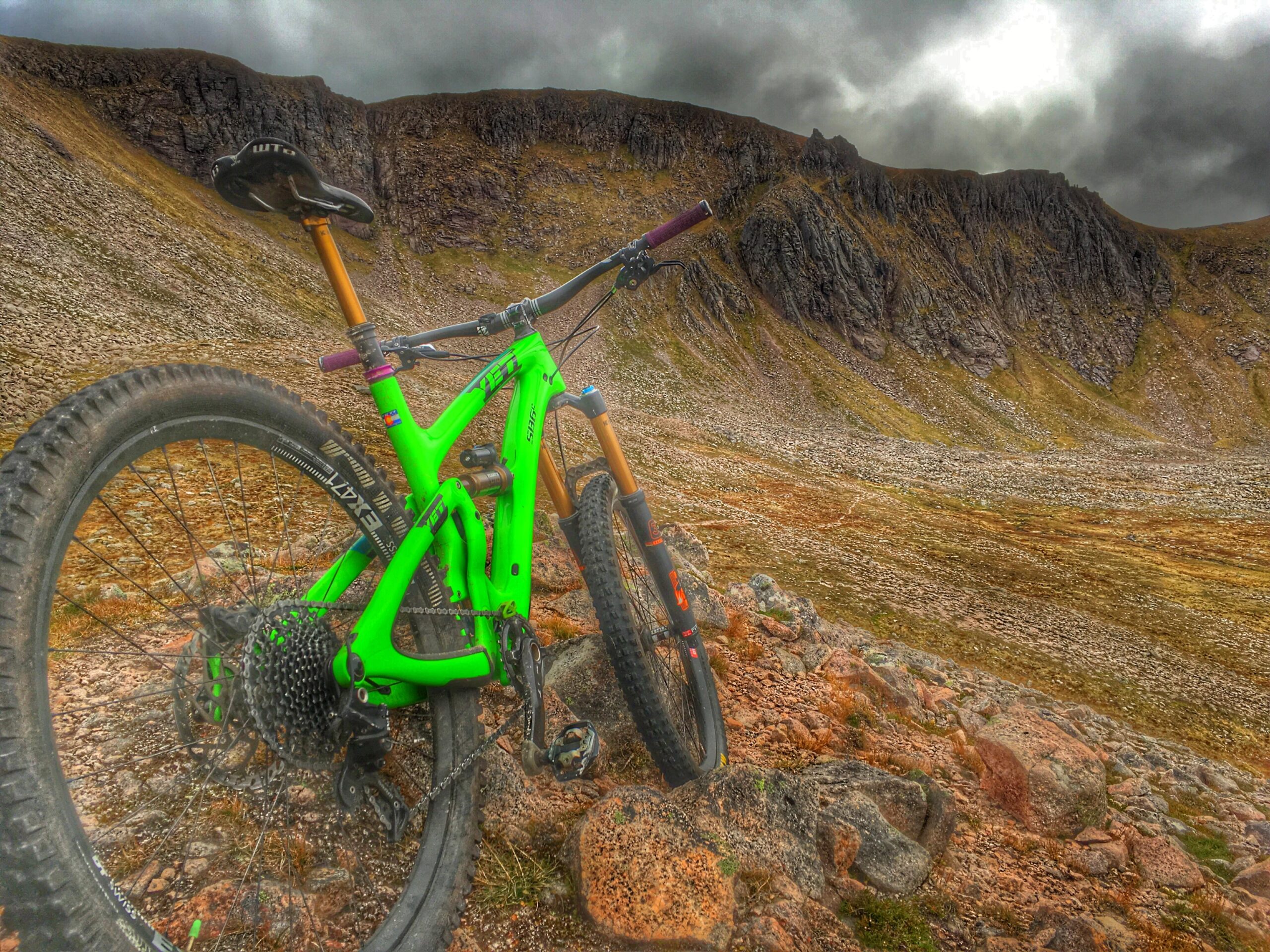 Yeti SB6: A bright green mountain bike leaning against a rock on a rugged, rocky hillside. In the background, a dramatic landscape with steep cliffs and cloudy skies, showcasing a mix of grass and rocky terrain.