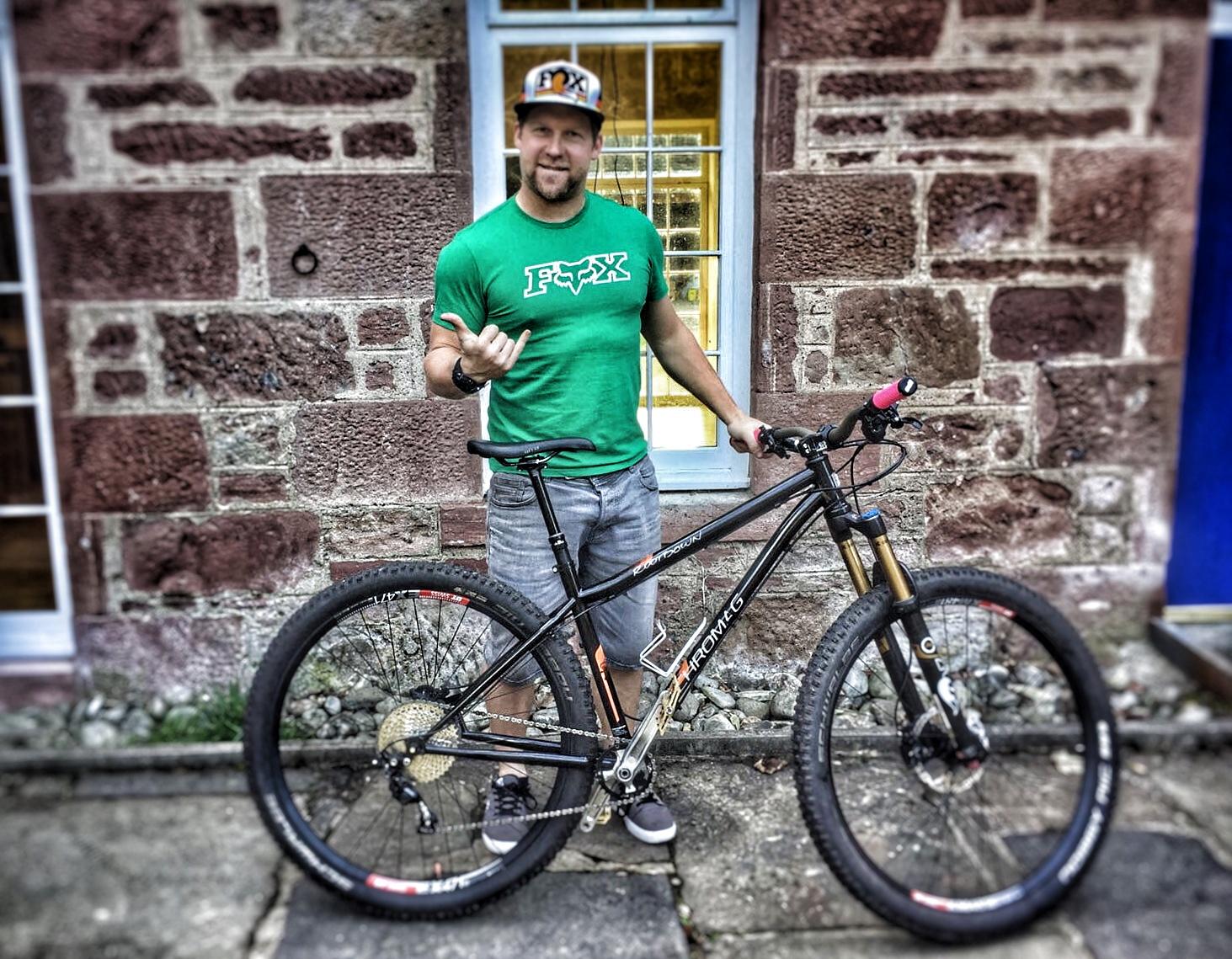 Chromag Rootdown: A man standing next to a mountain bike, wearing a green t-shirt and a cap. He is holding a thumbs-up gesture, with a stone wall and a blue door in the background. The bike features thick tires and a black frame.