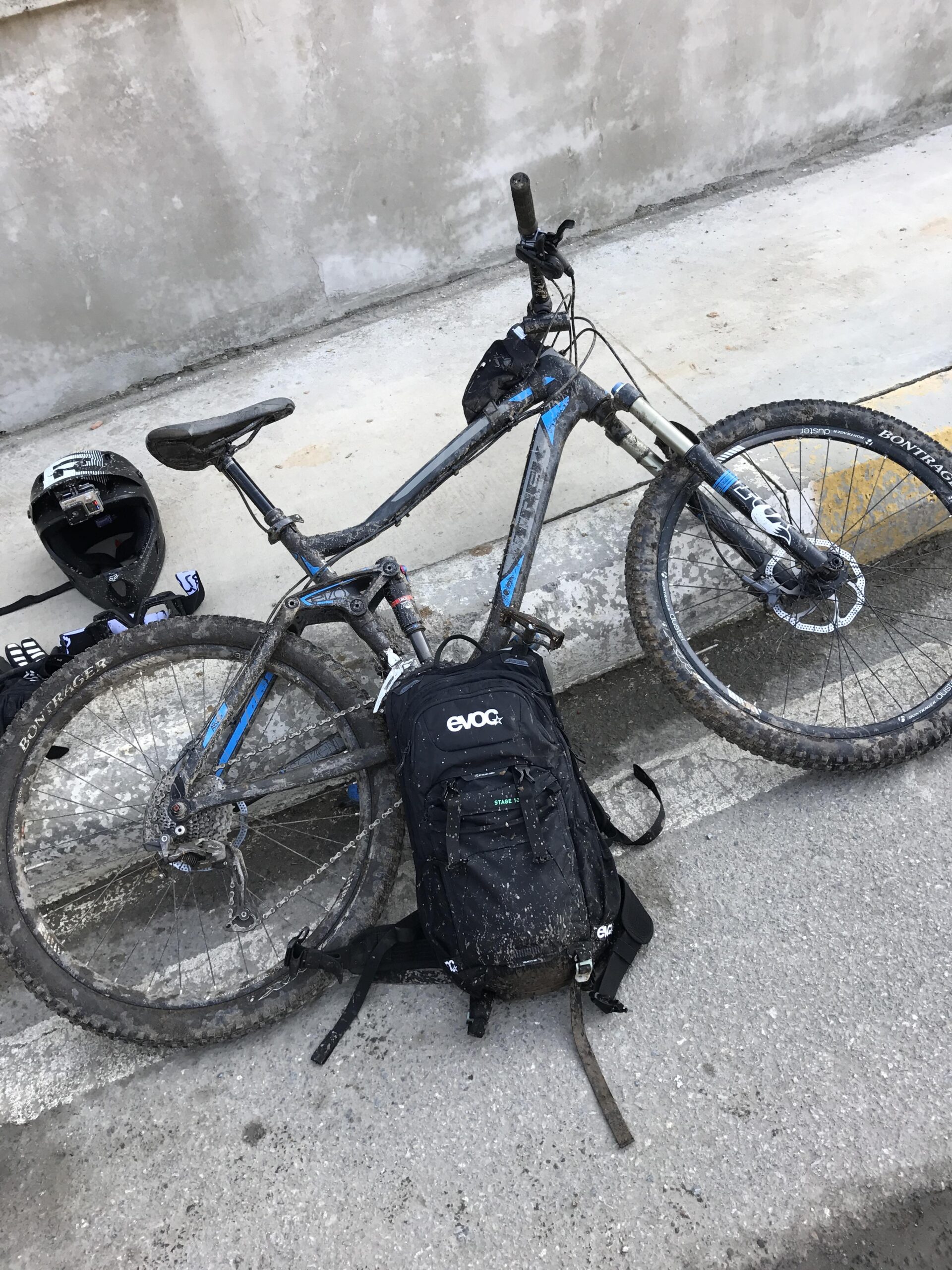 Trek Fuel EX 7: A muddy bicycle leaning against a concrete wall, accompanied by a black backpack labeled "evoc" and a black helmet. The bike features blue accents and appears to have been used on rough terrain, showing signs of dirt and wear.