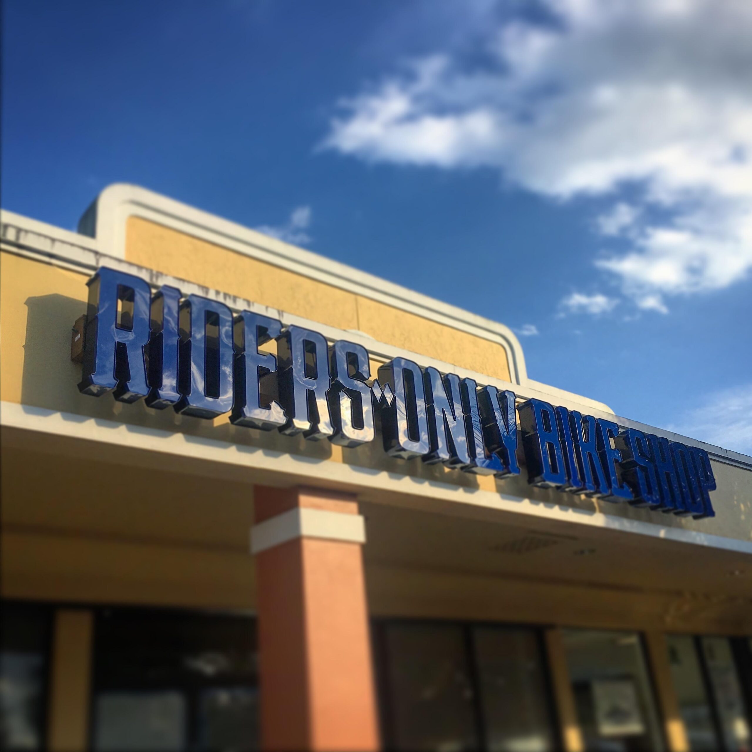 Signage of "Riders Only Bike Shop" displayed on the exterior of a building, under a clear blue sky with a few clouds visible.