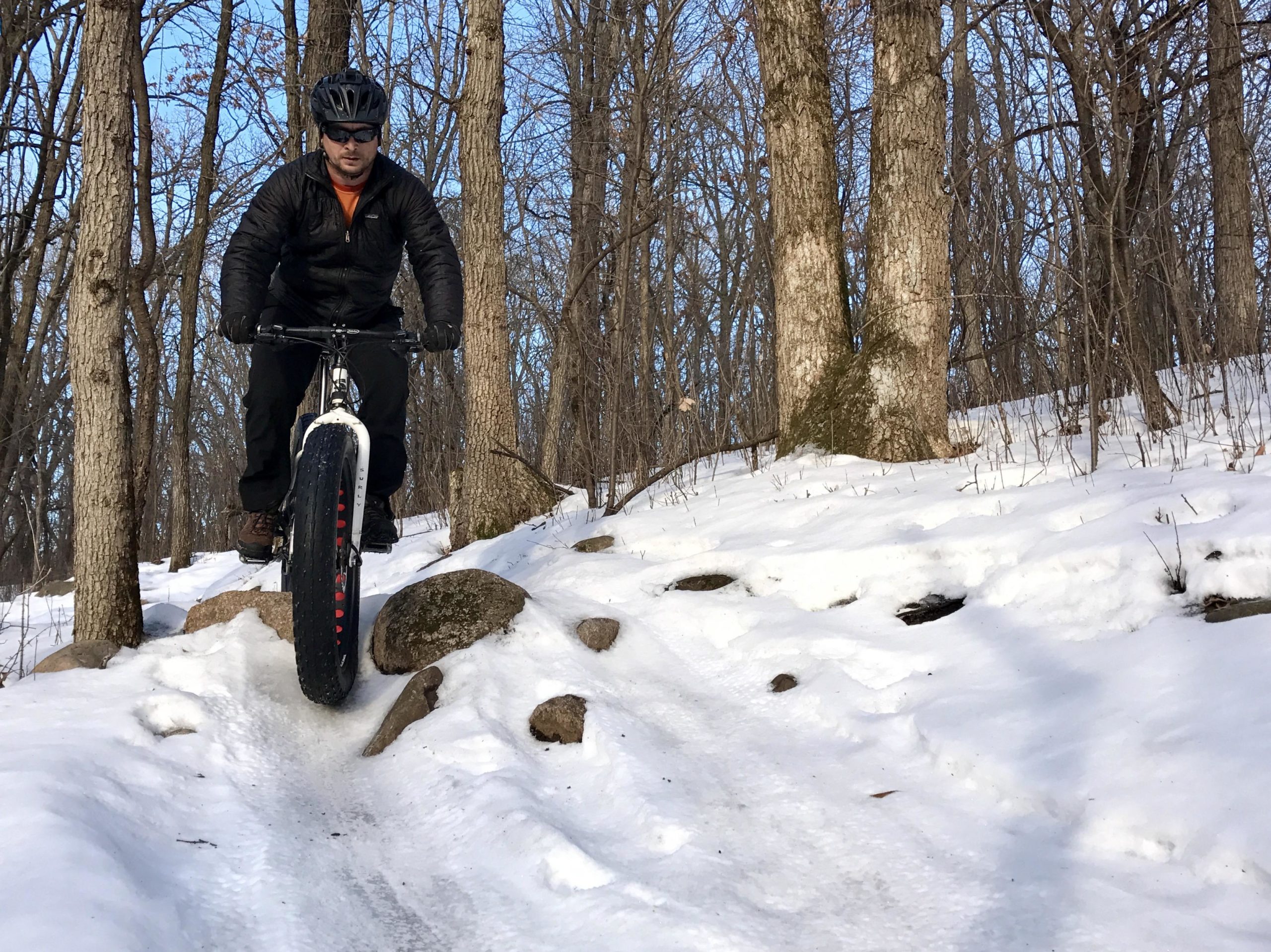 A person riding a fat bicycle on a snowy trail in a wooded area, navigating over rocks. The rider is wearing a helmet and dark clothing, with trees surrounding the path and a clear blue sky in the background. Lebanon Hills mountain bike trail.