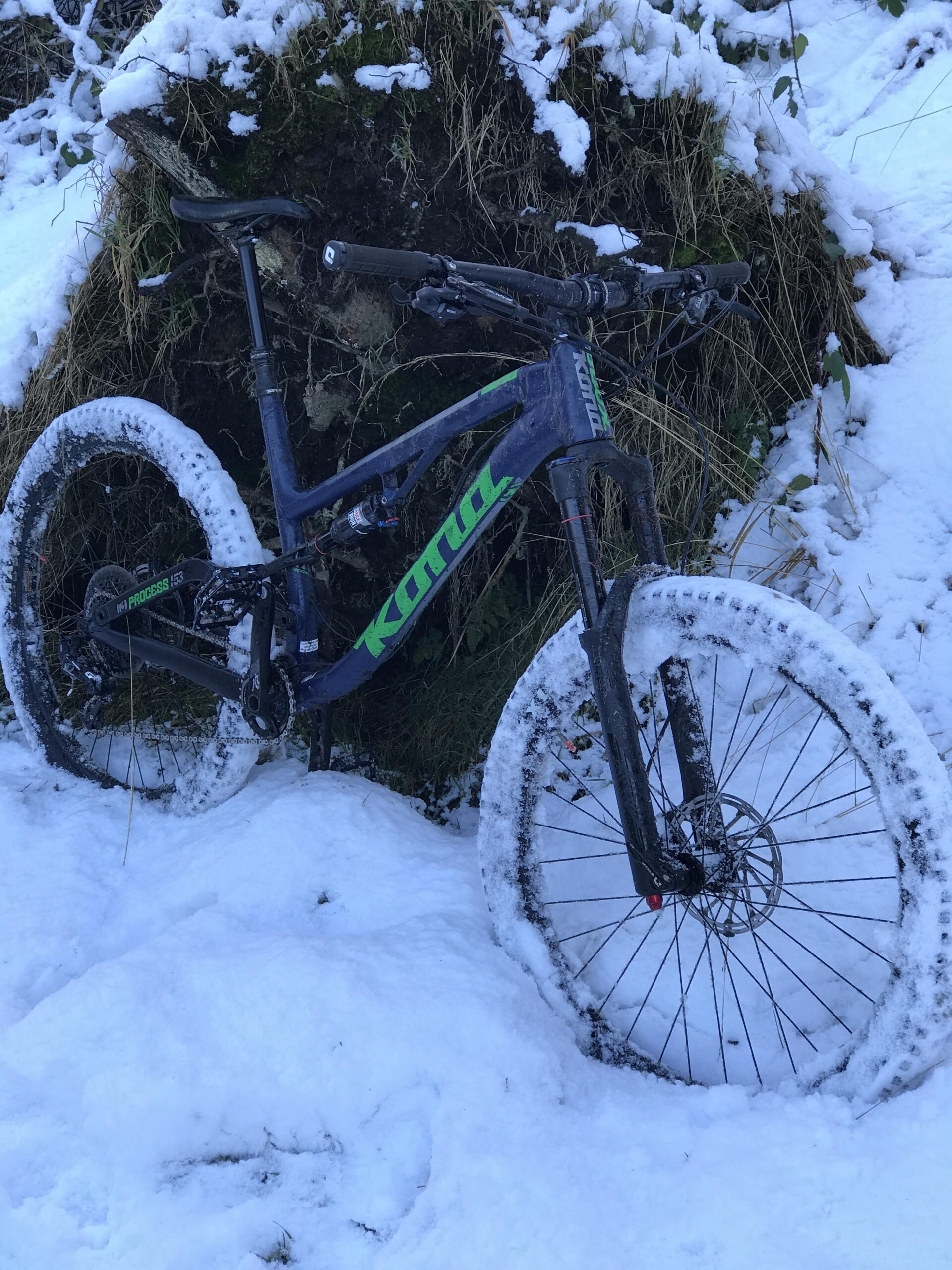 Kona Process 153: A mountain bike with a dark blue frame and green accents is leaning against a snow-covered bank. The bike's tires are partially coated in snow, indicating it has been used in winter conditions. Surrounding vegetation is also blanketed in a light layer of snow, creating a wintry scene.