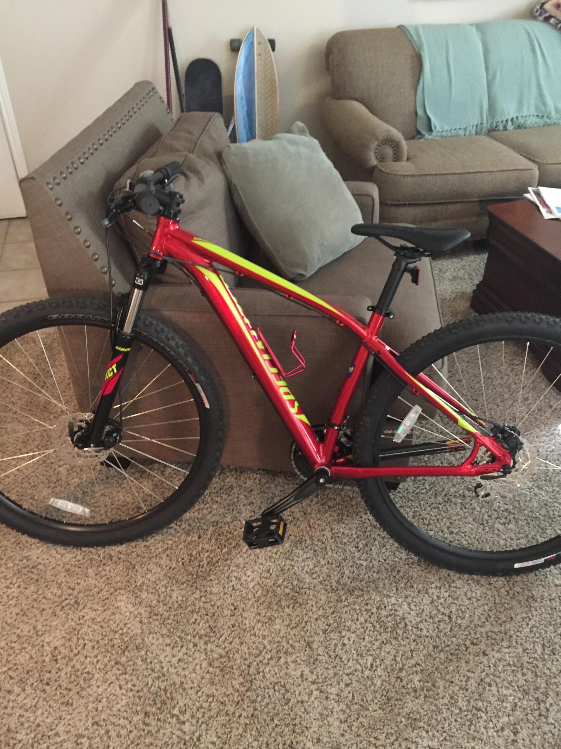 Specialized Rockhopper 29: A red and yellow mountain bike leaning against a couch in a living room, with a textured brown carpet and a coffee table nearby. Two gray cushions are on the couch, and a light blue blanket is draped over one side. In the background, there are two skateboards propped against the wall.