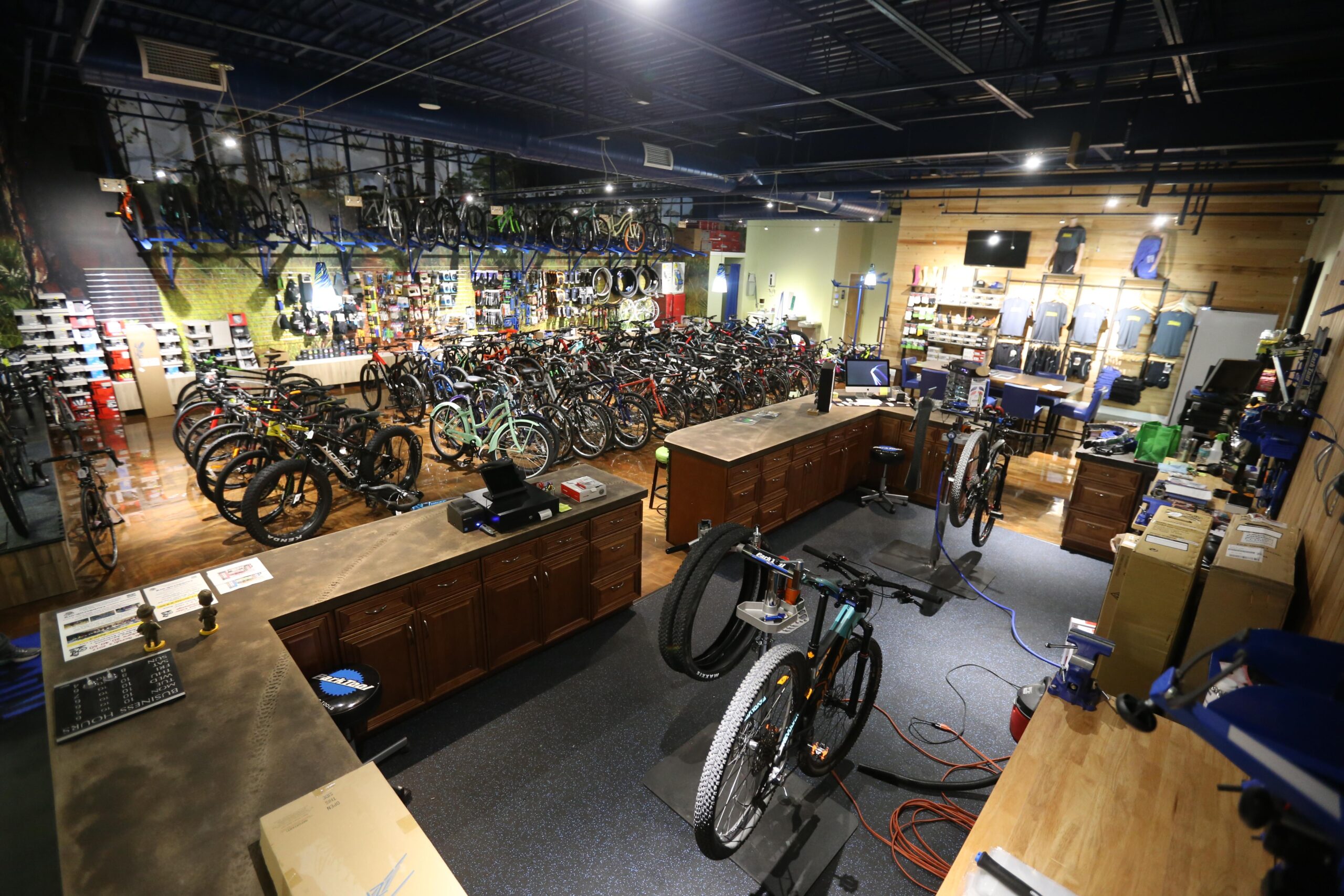A spacious bicycle shop interior featuring numerous bicycles displayed throughout the store. The scene includes a service area with repair stations, a wooden counter with registers, and various bike accessories on the walls. Bright lighting highlights the assortment of bikes, with a mix of mountain and road bikes visible.