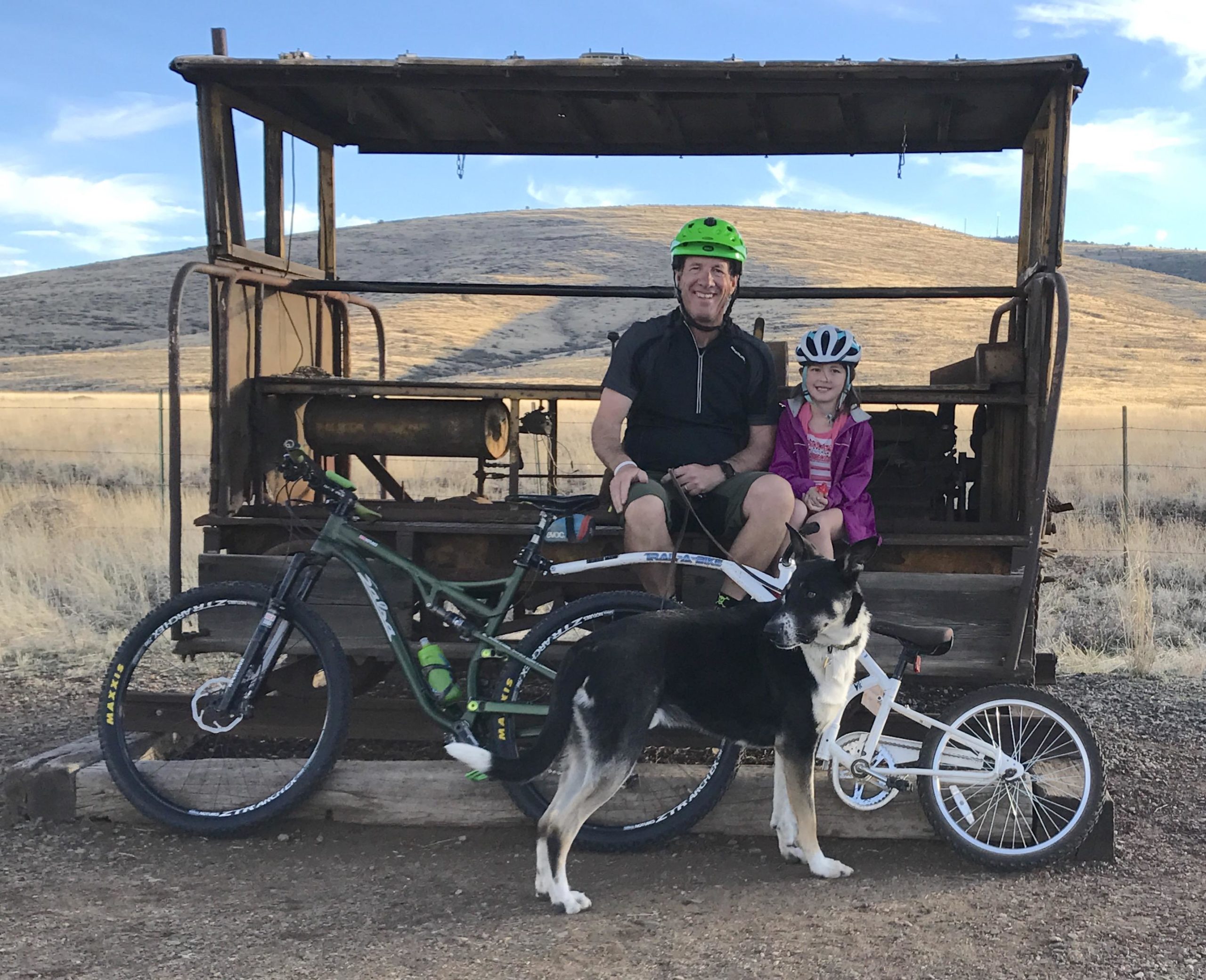 A man and a young girl, both wearing helmets, are sitting on an old metal structure in an open field. The man is smiling next to a mountain bike, while the girl is holding onto a small white bicycle. A black and white dog stands near them, with hills in the background and a clear sky above. The scene captures a moment of outdoor adventure. Prescott Peavine / Iron King mountain bike trail.