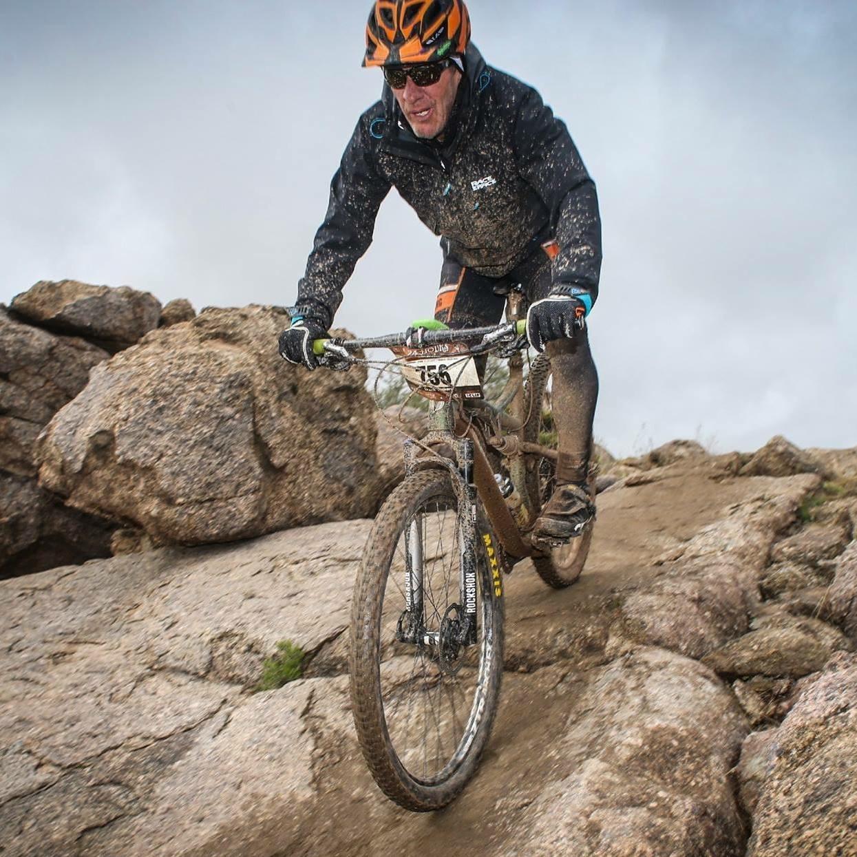 A mountain biker navigating a rocky terrain, covered in dirt and mud, wearing a helmet and sunglasses. The biker is leaning forward, concentrating on maintaining balance while riding downhill amidst a cloudy sky. 24 Hour Course mountain bike trail.