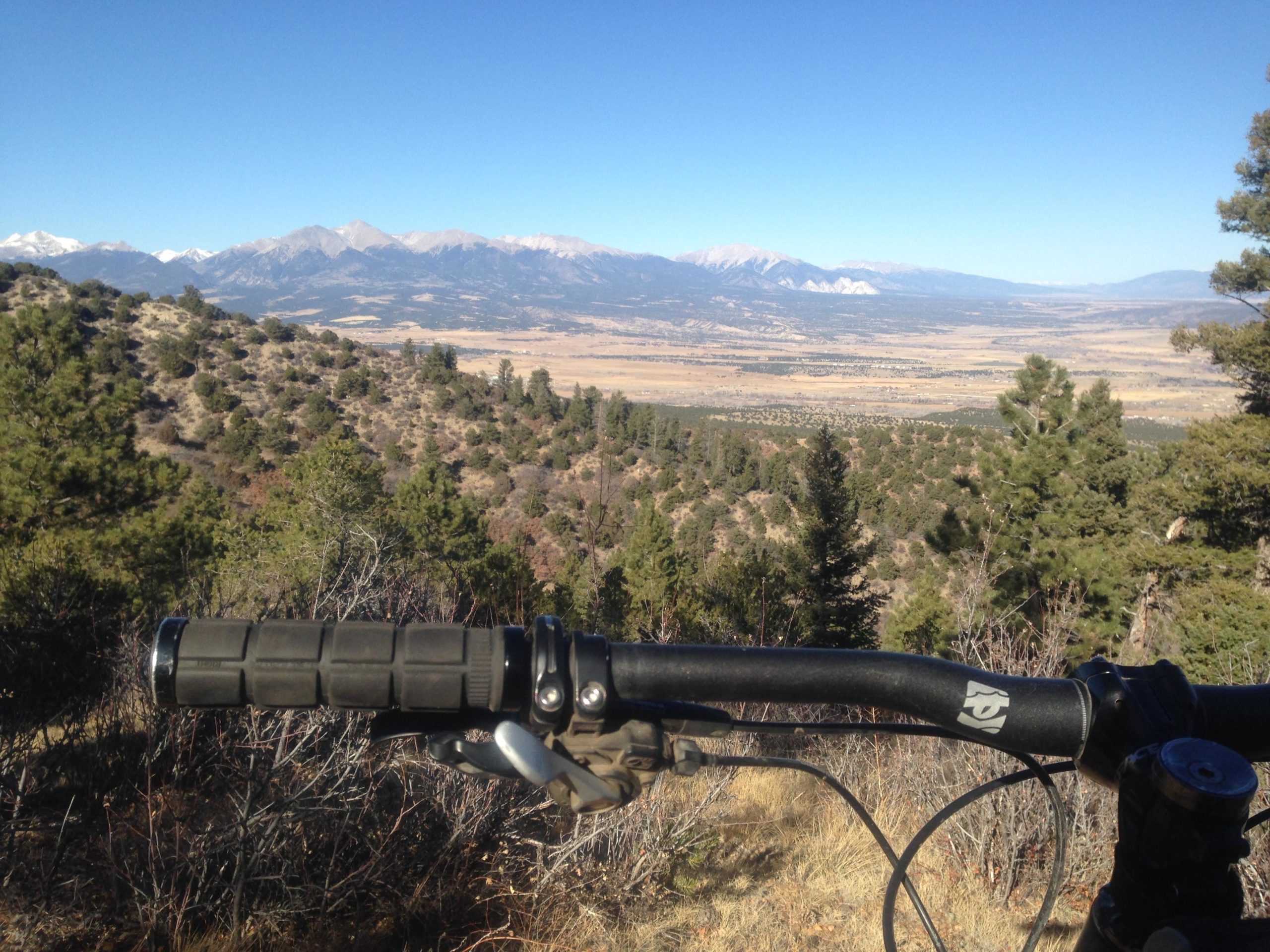 A mountain bike handlebar in the foreground, overlooking a scenic view of rolling hills and distant snow-capped mountains under a clear blue sky. The landscape includes scattered trees and open fields, showcasing the natural beauty of the area. Rainbow Trail: Methodist Mountain Thd to Bear Creek Thd mountain bike trail.