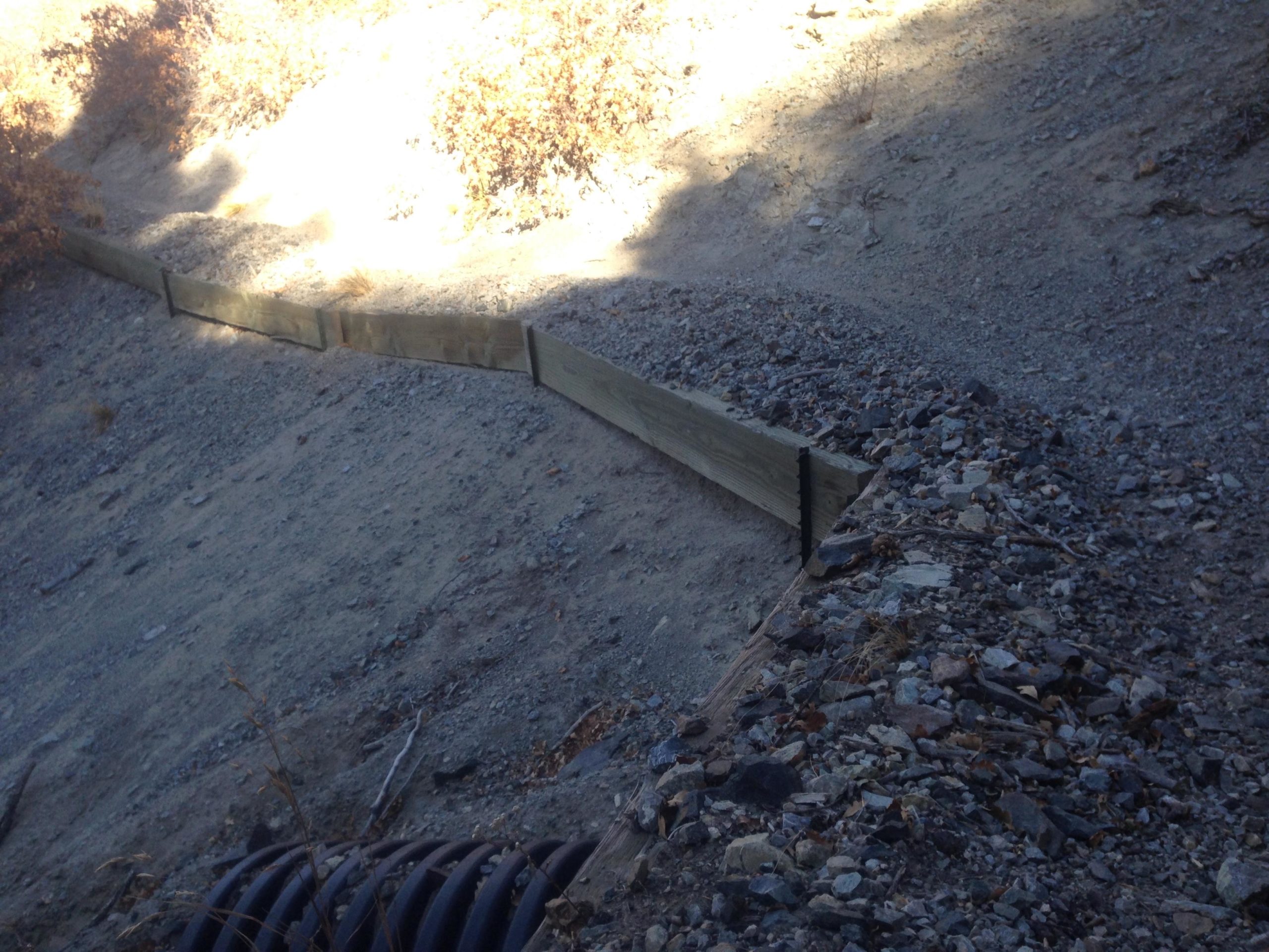 A sloped, rocky terrain featuring a wooden barrier along the edge, with gravel and sparse vegetation visible in the background. Sunlight filters through, illuminating parts of the landscape and creating shadows. Rainbow Trail: Methodist Mountain Thd to Bear Creek Thd mountain bike trail.