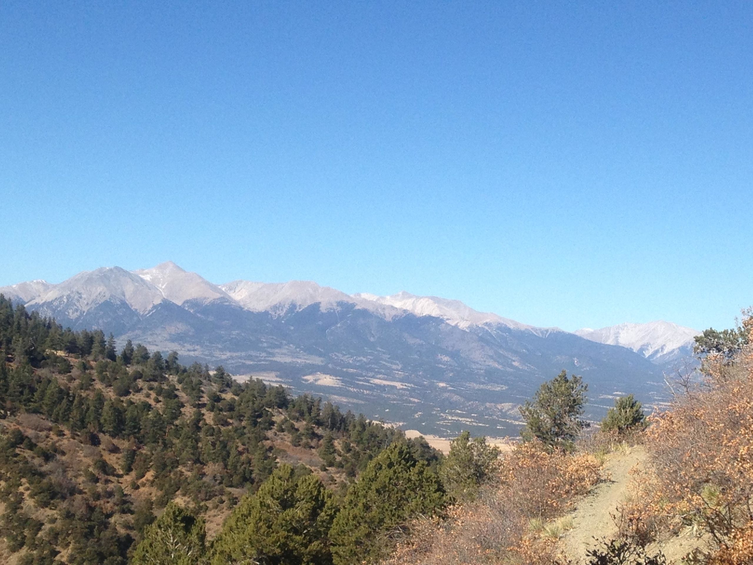 A scenic view of a mountain range under a clear blue sky, with snow-dusted peaks and dense evergreen forests in the foreground. The landscape features rolling hills and valleys, creating a tranquil natural setting. Rainbow Trail: Methodist Mountain Thd to Bear Creek Thd mountain bike trail.