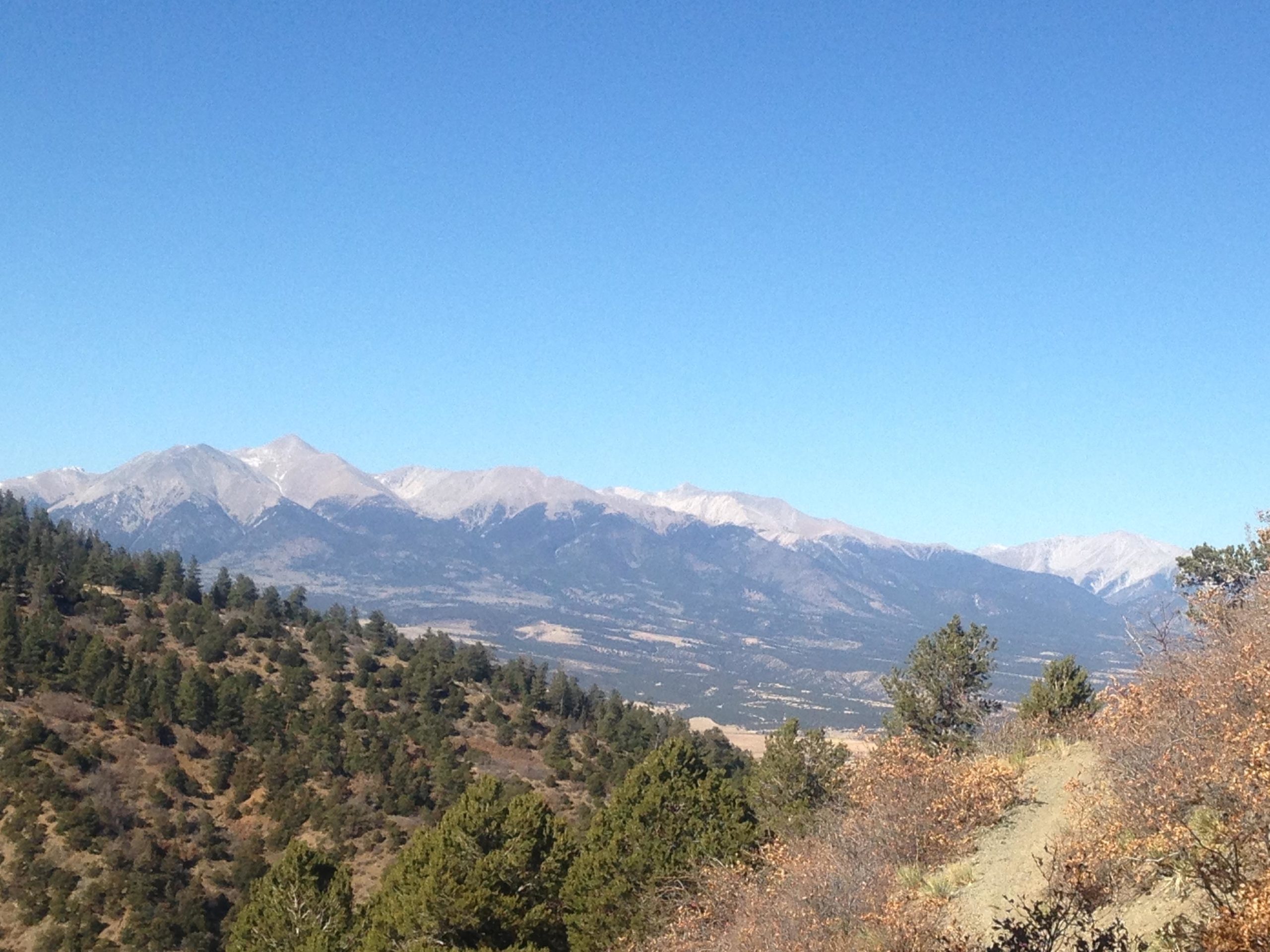 A panoramic view of a mountainous landscape under a clear blue sky, featuring snow-capped peaks in the distance and lush green forests in the foreground. Rainbow Trail: Methodist Mountain Thd to Bear Creek Thd mountain bike trail.