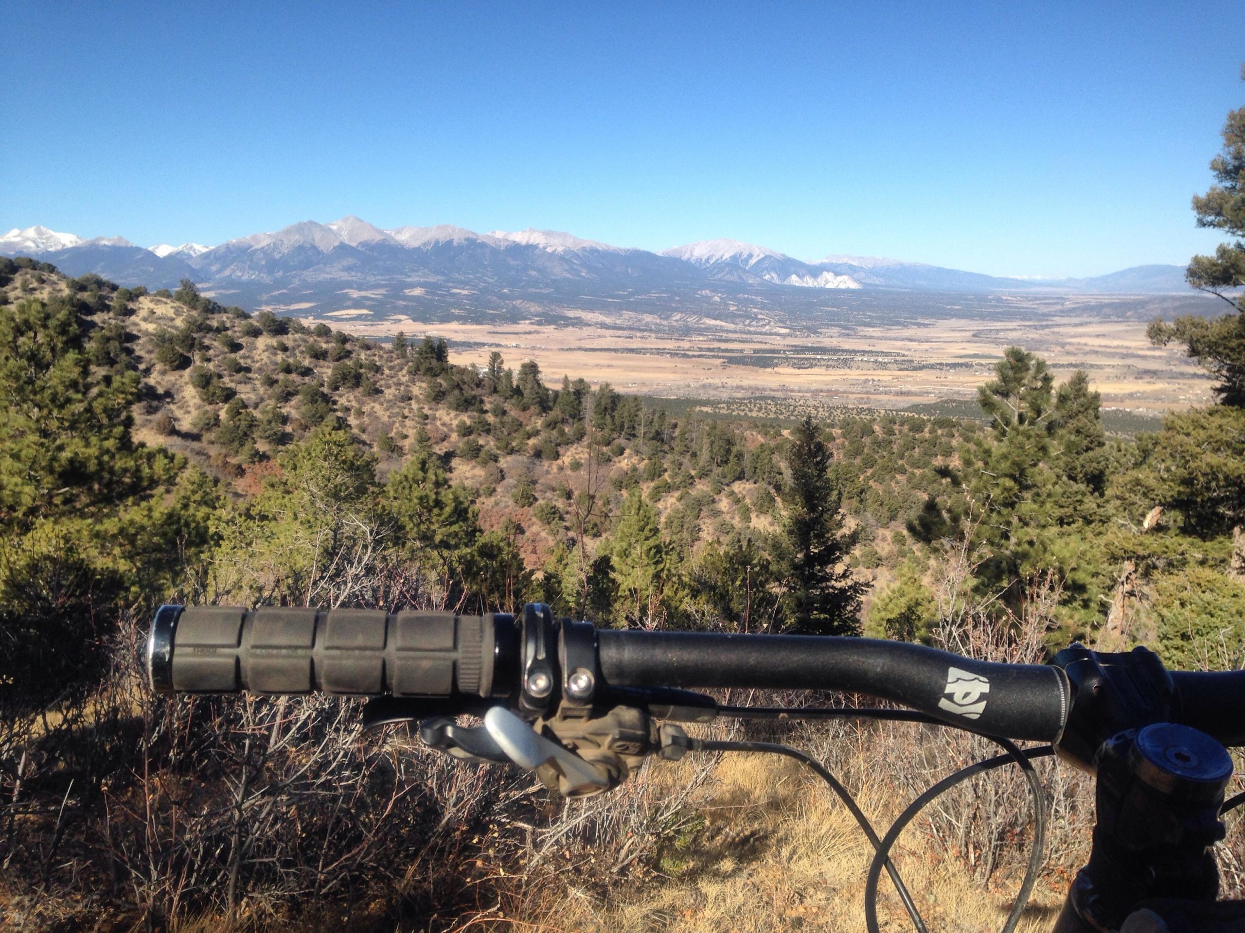 A mountain bike handlebar is prominently featured in the foreground, with a breathtaking view of snow-capped mountains and a lush valley stretching out in the background under a clear blue sky. The scene captures the essence of outdoor adventure and natural beauty. Rainbow Trail: Methodist Mountain Thd to Bear Creek Thd mountain bike trail.