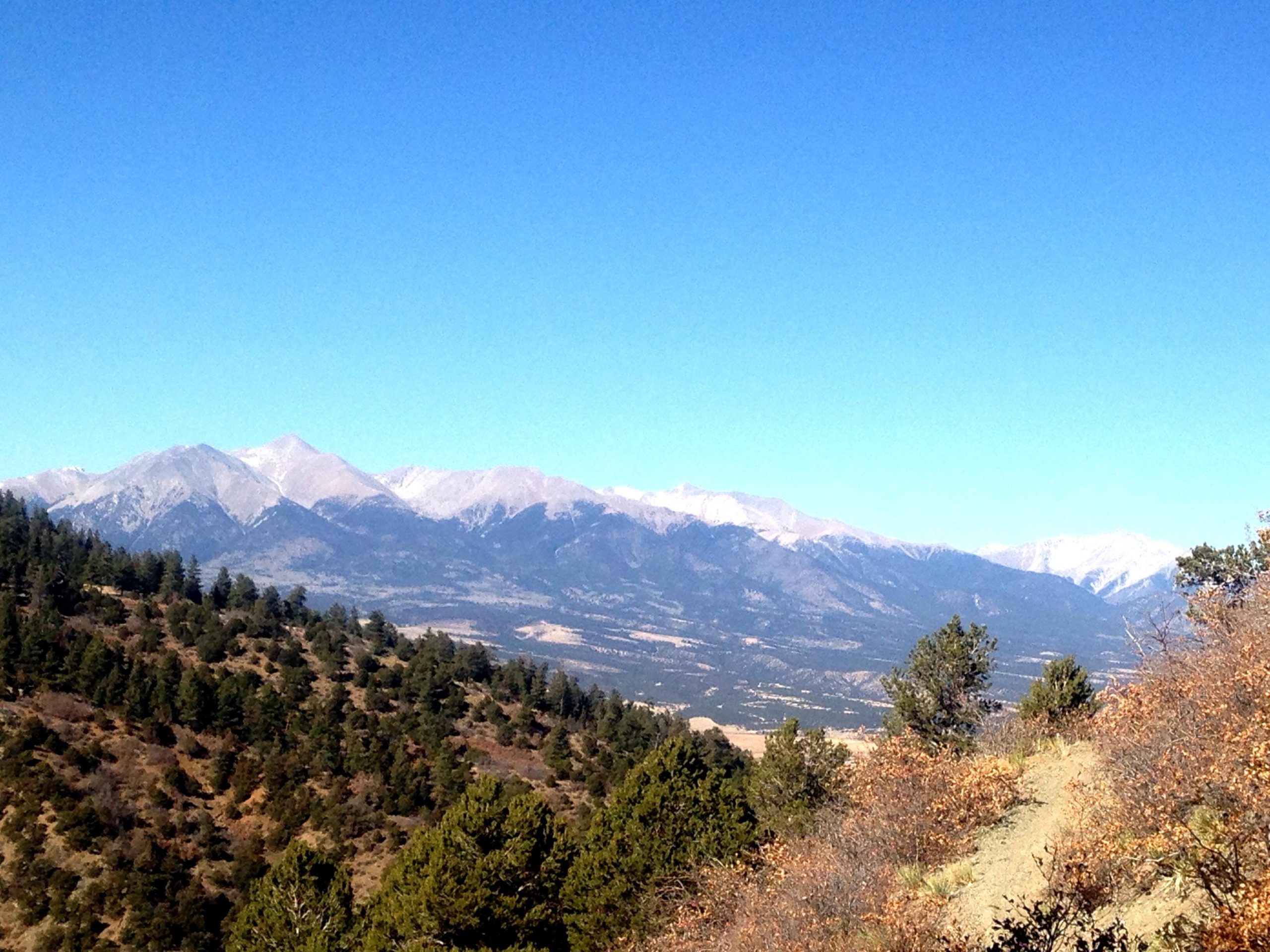 A landscape view of mountains with snow-capped peaks under a clear blue sky, surrounded by rolling hills and patches of evergreen trees in the foreground. Rainbow Trail: Methodist Mountain Thd to Bear Creek Thd mountain bike trail.