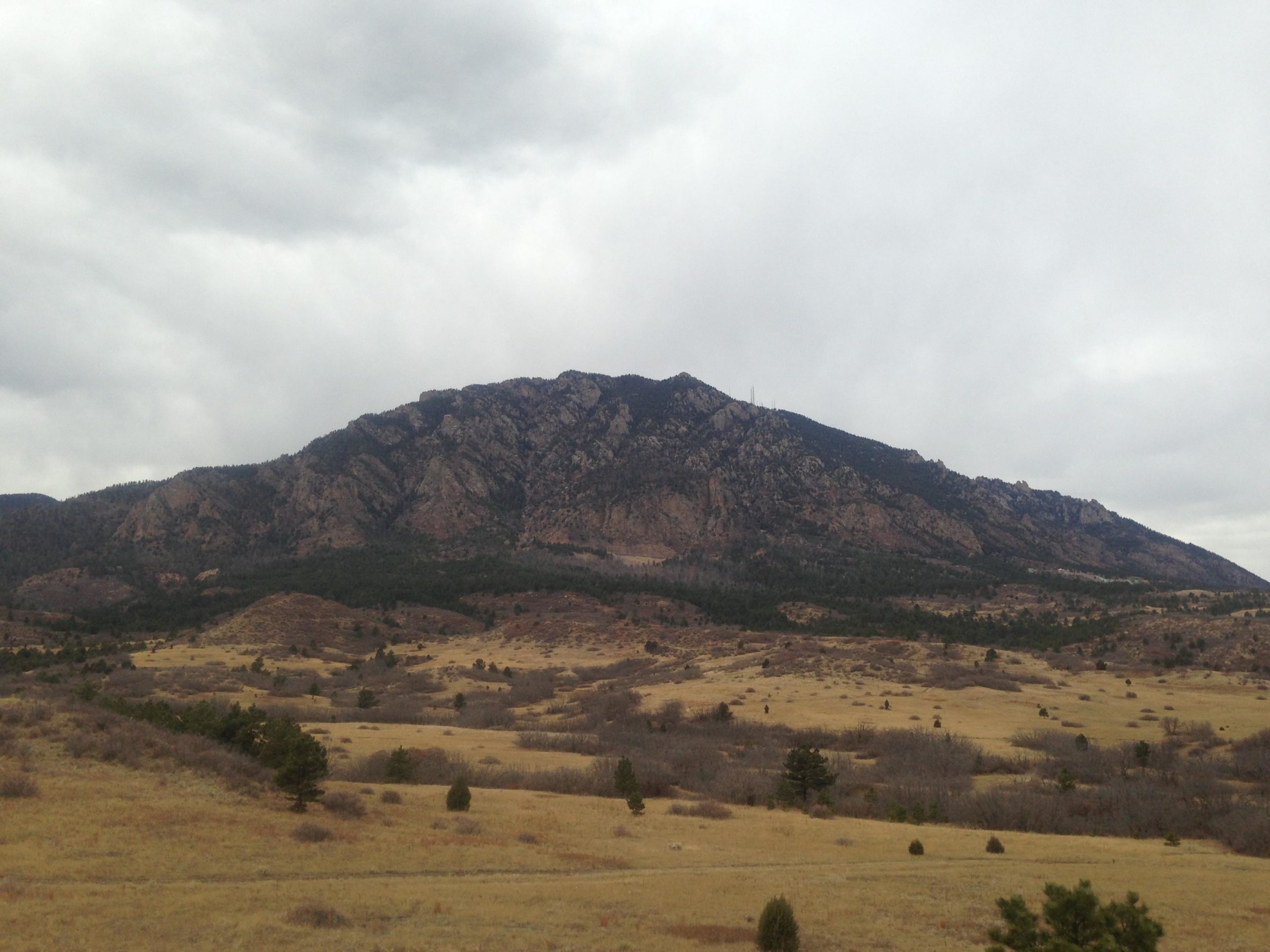 A panoramic view of a rugged mountain range under a cloudy sky, surrounded by open grassland and patches of trees. The mountain features rocky outcrops and is partially covered with greenery, creating a stark contrast with the golden hues of the plains below. Cheyenne Mountain State Park mountain bike trail.