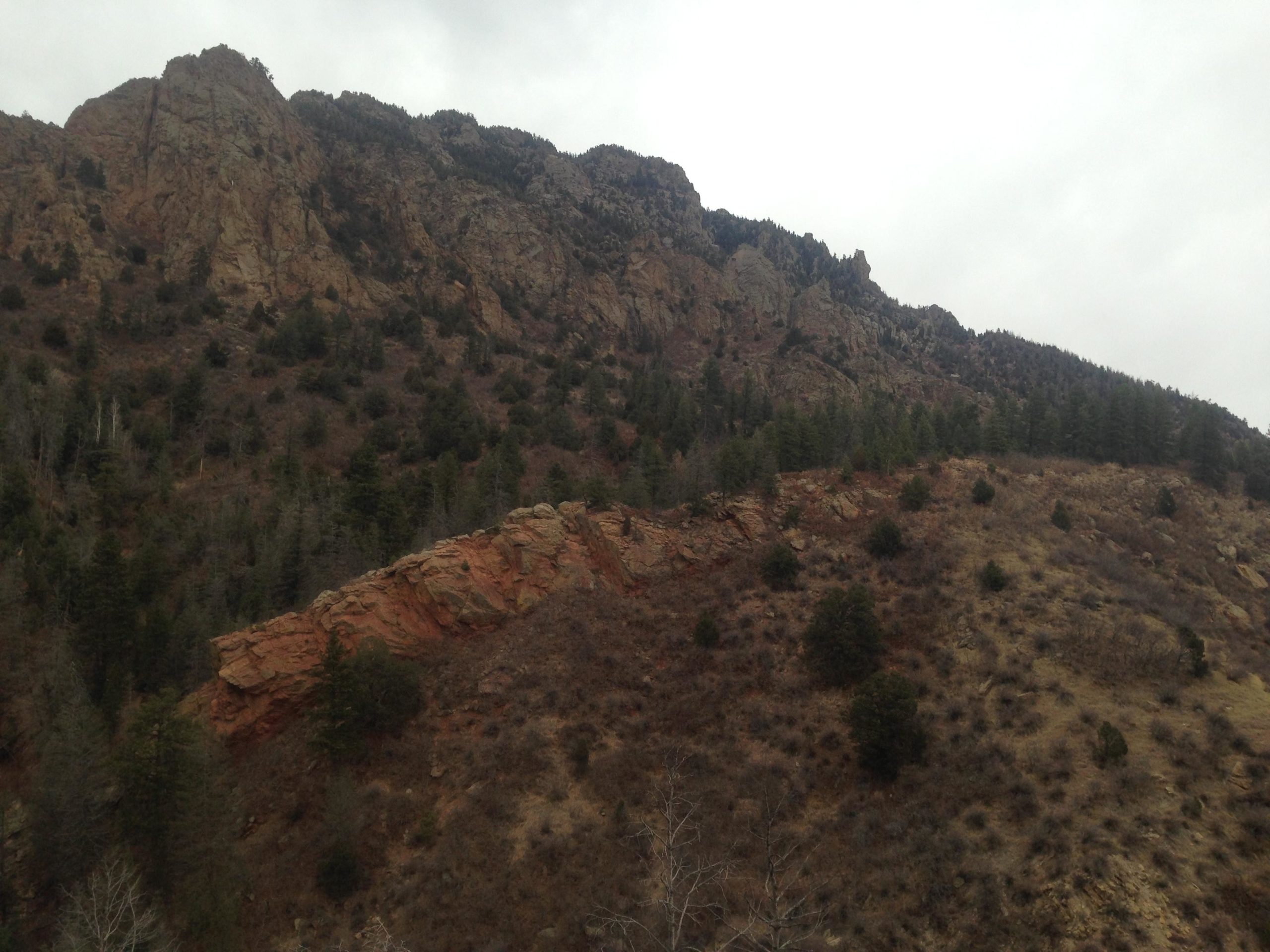 A rugged mountain landscape featuring steep, rocky cliffs topped with evergreen trees, sloping down to a lower hillside with scattered vegetation. The sky is overcast, creating a moody atmosphere. Cheyenne Mountain State Park mountain bike trail.