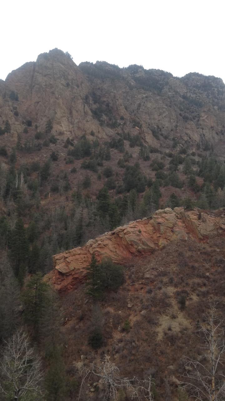 A rugged mountain landscape featuring steep, rocky cliffs and scattered evergreen trees. The terrain includes a prominent red rock formation, with patches of dry, brown vegetation. The sky is overcast, adding a dramatic atmosphere to the scene. Cheyenne Mountain State Park mountain bike trail.