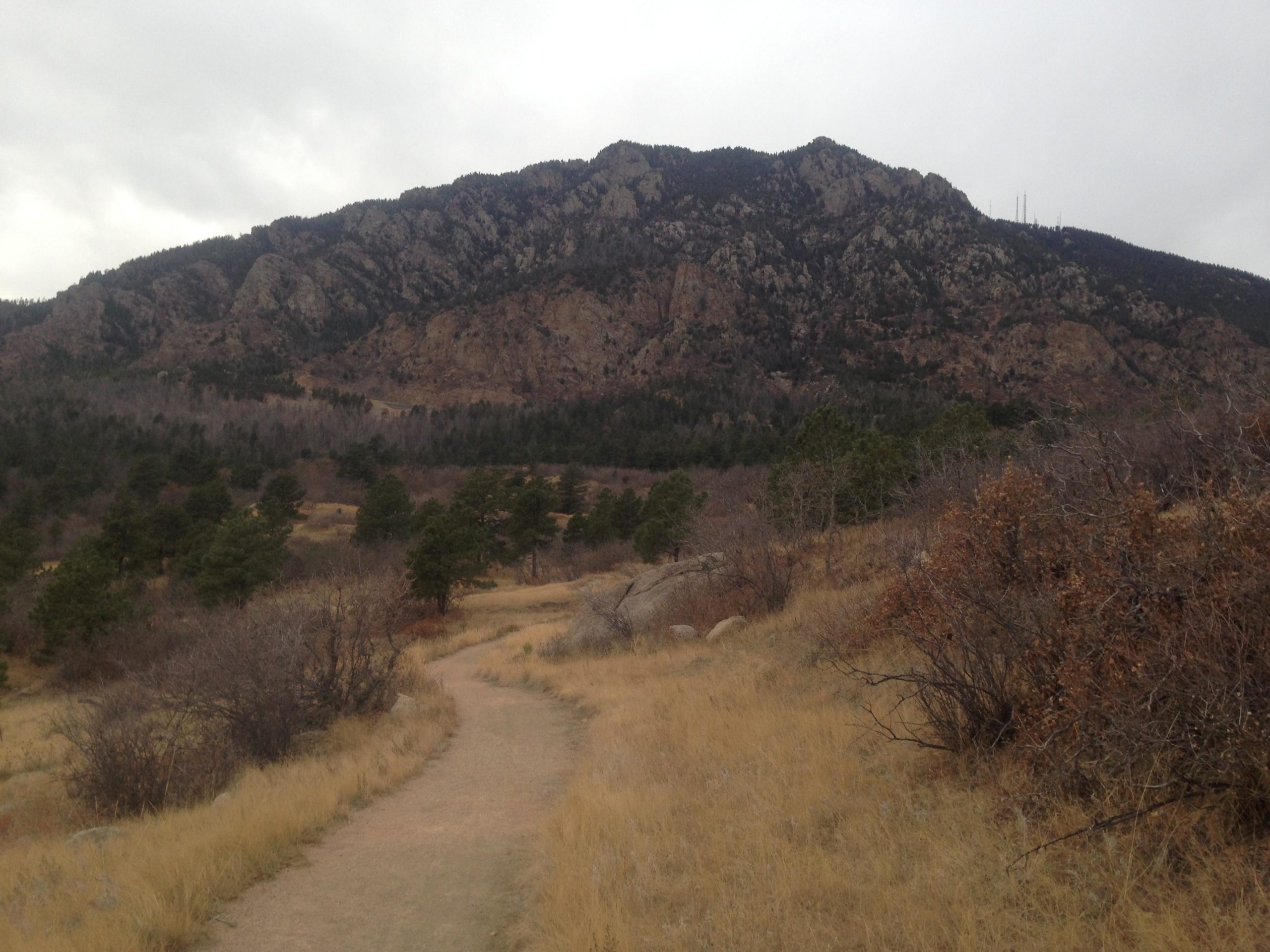 A winding dirt path leads through dry grassland and sparse trees towards a rocky mountain in the background, under a cloudy sky. Cheyenne Mountain State Park mountain bike trail.