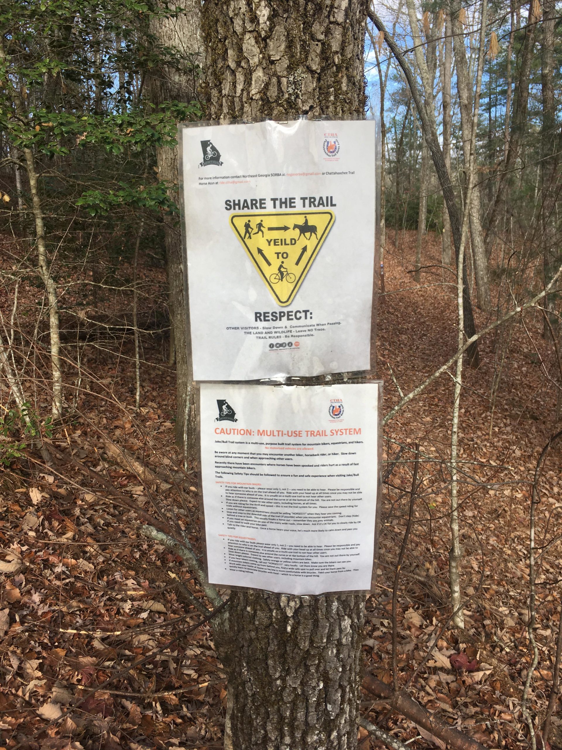 Two informational signs affixed to a tree in a wooded area. The top sign features a yellow triangle symbol instructing trail users to "Share the Trail" and "Yield to" pedestrians and cyclists. The bottom sign provides cautionary details about the multi-use trail system, emphasizing safety guidelines and respect for all trail users. The ground is covered in fallen leaves, and trees surround the area. Jake to Bull Mountain Connecter mountain bike trail.