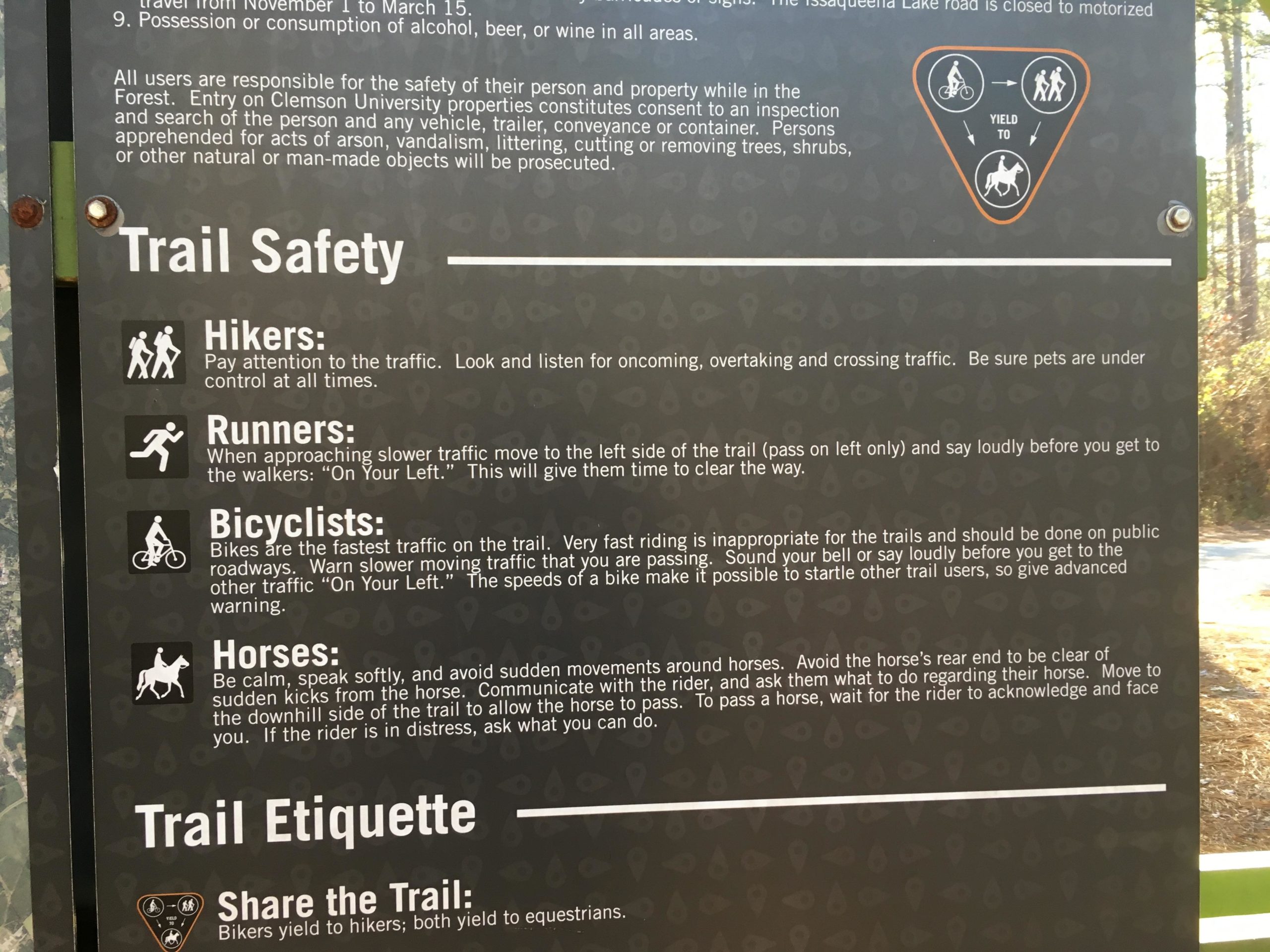 Signage detailing trail safety and etiquette rules for hikers, runners, bicyclists, and horseback riders. The sign includes specific instructions for each group on how to navigate the trails safely and courteously, such as yielding to hikers, announcing oneself when passing, and being cautious around horses. Issaqueena Lake mountain bike trail.