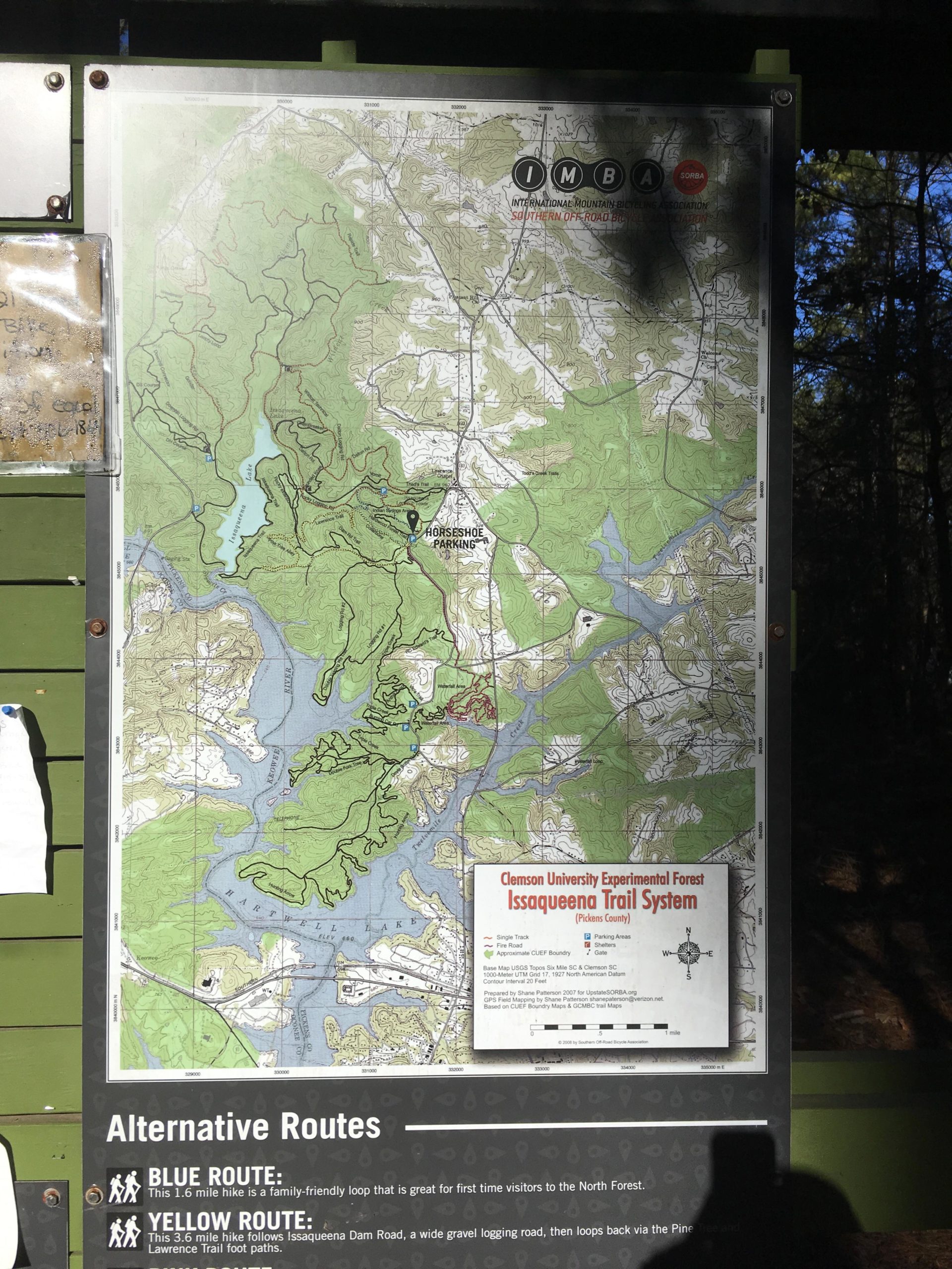 A map of the Issaqueena Trail System located in the Clemson University Experimental Forest, showing various hiking routes, including the Blue Route and Yellow Route, along with features like parking areas and landmarks. The map includes topographical details and is situated on a wooden display board in a forested area. Issaqueena Lake mountain bike trail.