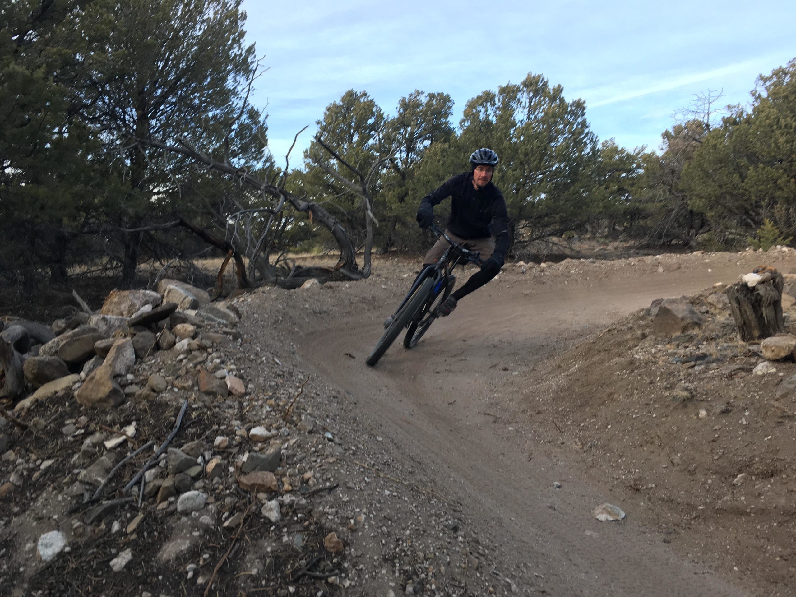 A mountain biker leans into a turn on a dirt trail surrounded by trees and rocky terrain. The rider is wearing a helmet and dark clothing, showcasing an energetic motion as they navigate the winding path. Chicken Dinner mountain bike trail.