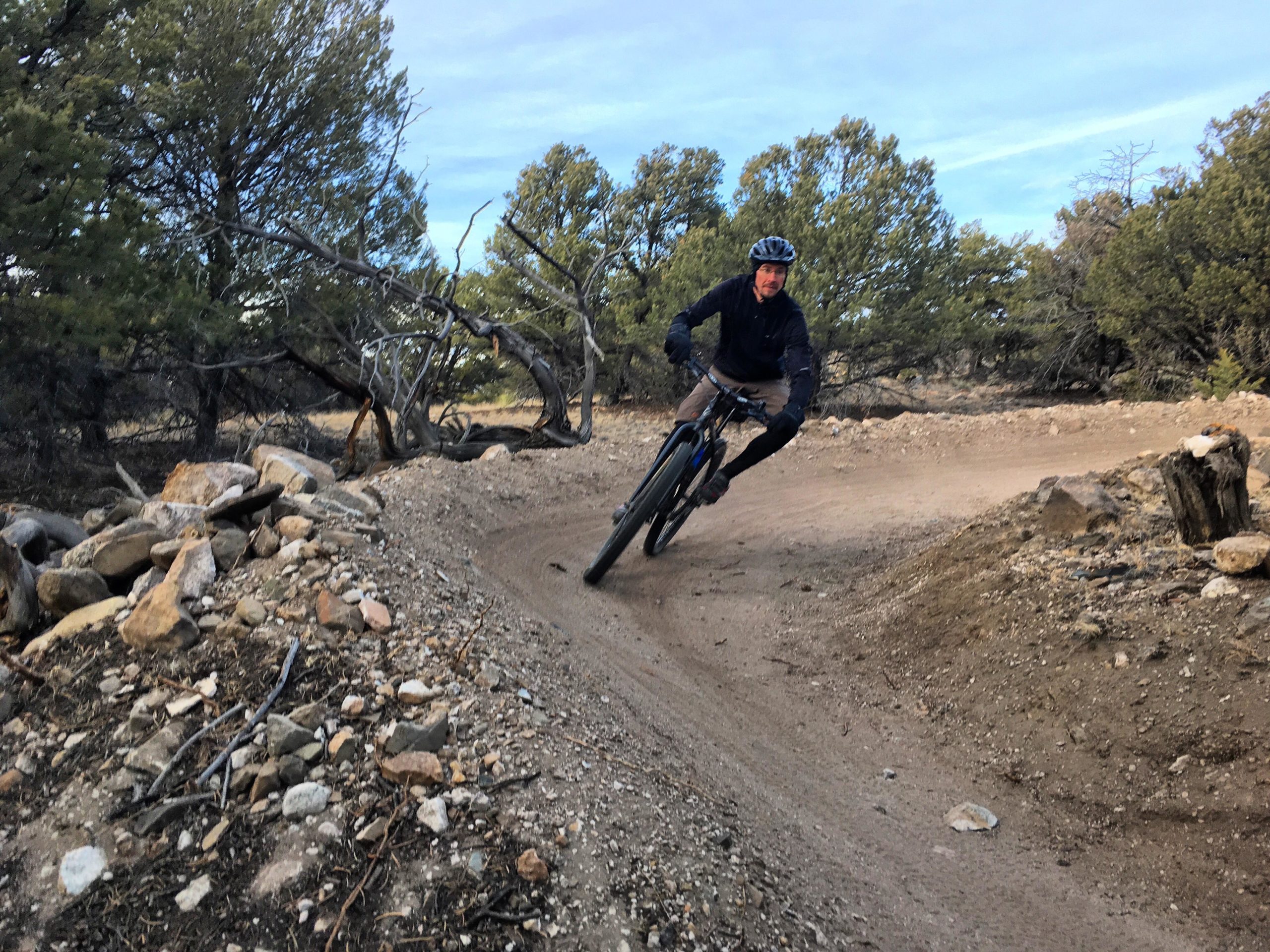A mountain biker riding on a dirt trail, leaning into a turn with trees and rocks in the background, under a clear blue sky. Chicken Dinner mountain bike trail.