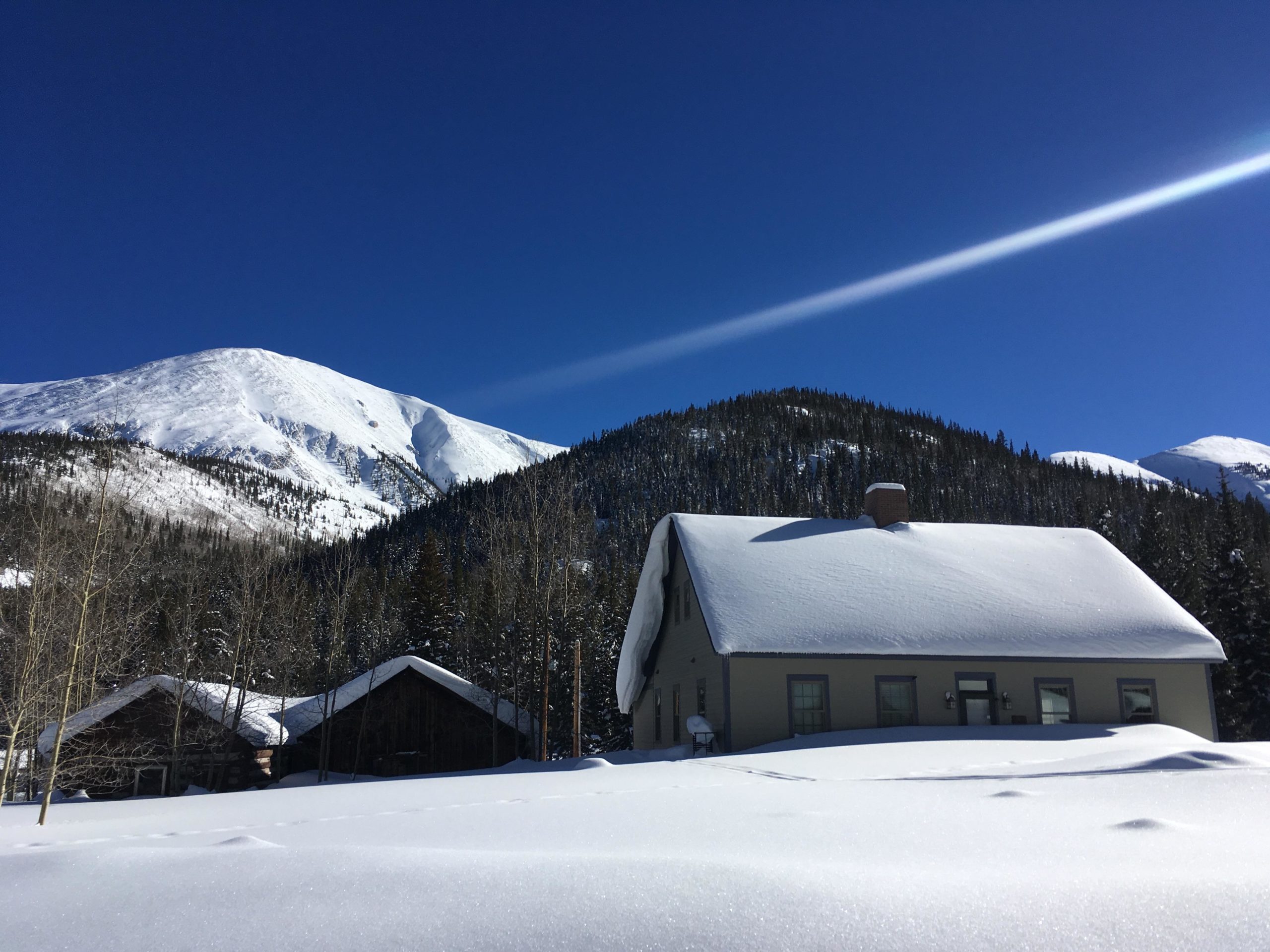 A snowy landscape with a house in the foreground, featuring a sloped roof blanketed in snow. In the background, tall mountains are covered in snow, beneath a clear blue sky. The scene conveys a peaceful winter setting with trees dotting the lower slopes of the mountains. Tincup Pass Road mountain bike trail.