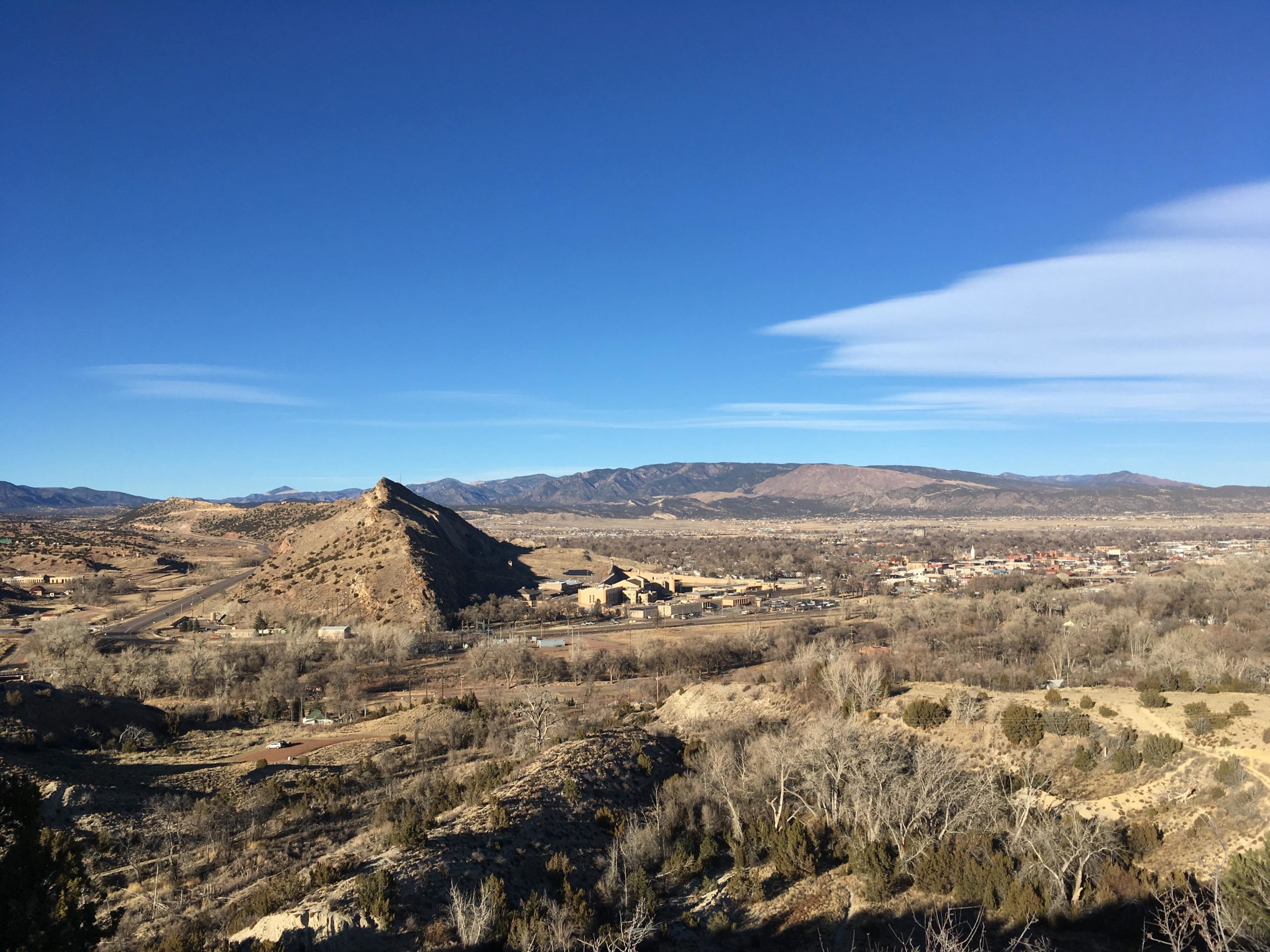 A panoramic view of a hilly landscape under a clear blue sky, featuring a prominent hill in the foreground with a triangular peak. The background showcases a range of mountains and a valley dotted with sparse vegetation, including trees and shrubs. Below, a small town can be seen nestled in the valley, with roads and buildings visible. The scene conveys a serene and natural setting. South Canon Trails mountain bike trail.