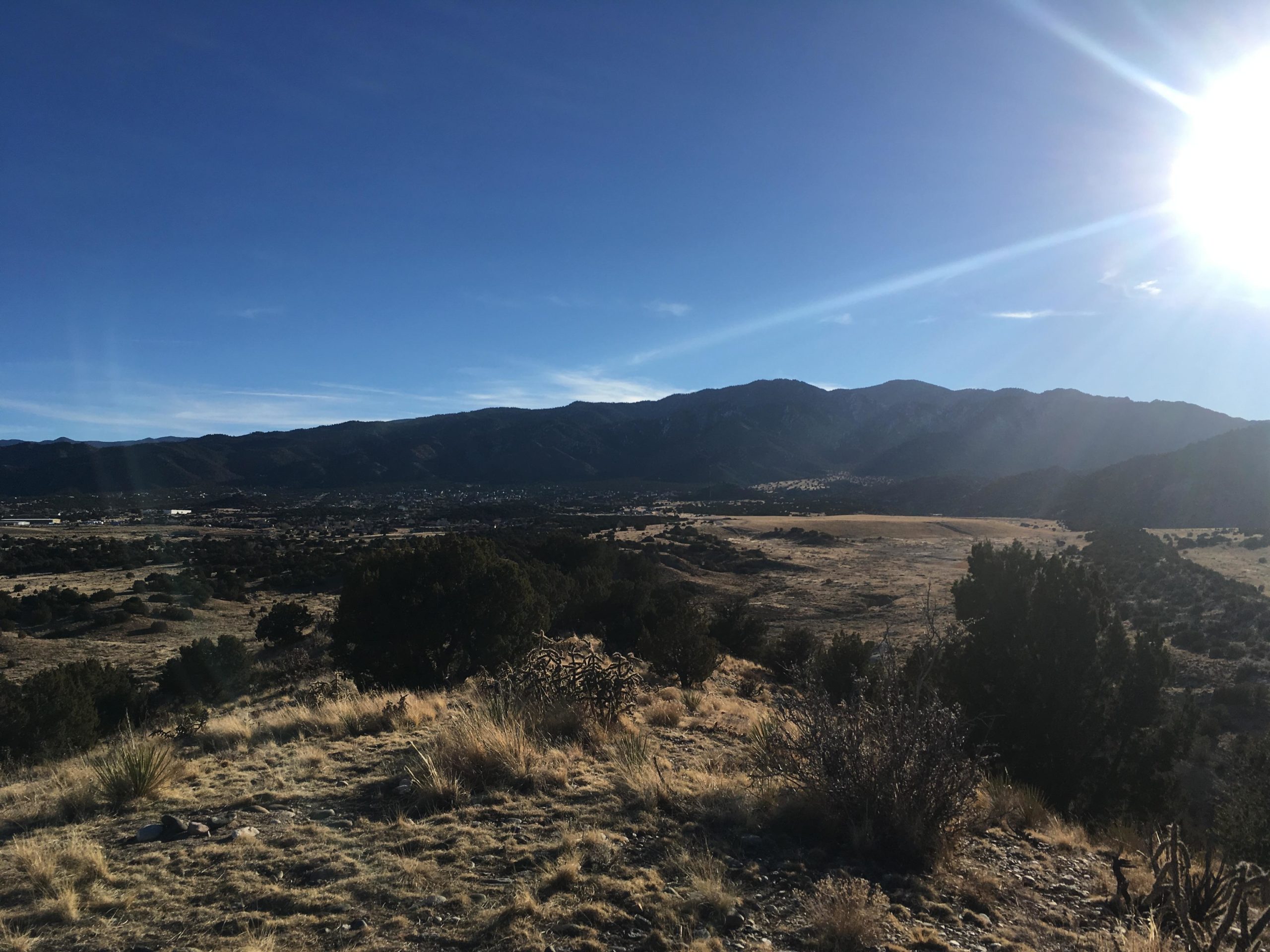 A scenic view of a mountainous landscape under a bright blue sky, with the sun shining in the top right corner. In the foreground, dry grass and sparse vegetation lead down to a valley, which is bordered by rolling hills and distant mountains. Light clouds streak the sky, indicating a clear day. South Canon Trails mountain bike trail.