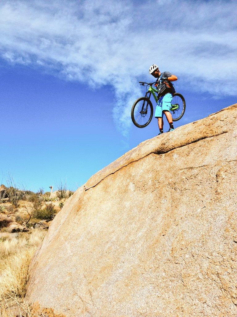 A young person standing on top of a large rock, holding a mountain bike, against a clear blue sky with wispy clouds. The landscape features dry vegetation typical of a desert environment. 50-year Trail / Golder Ranch mountain bike trail.