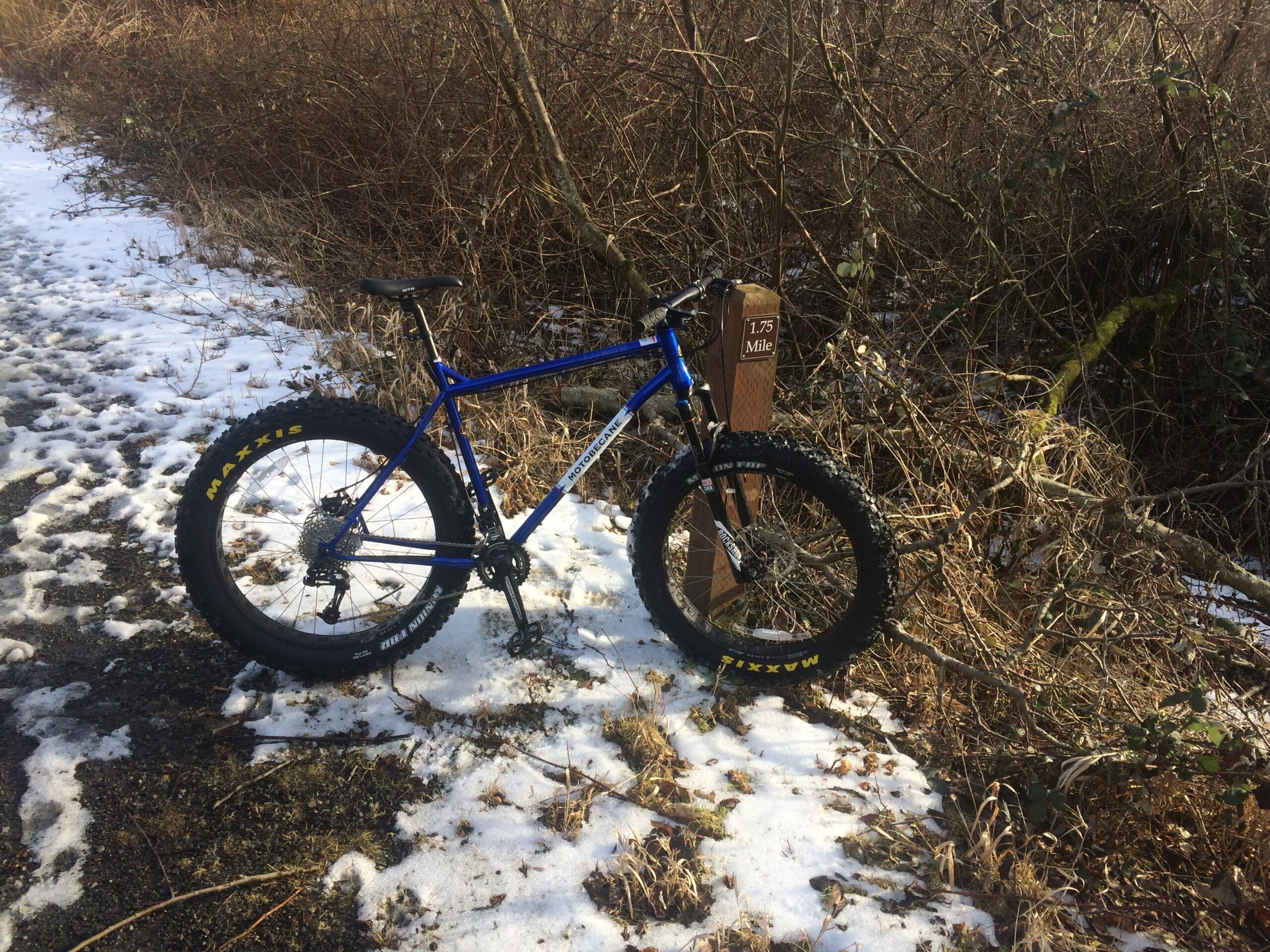 Motobecane Lurch: A blue fat bike parked on a snowy trail next to a wooden sign indicating "1.75 Mile." Surrounding scenery includes sparse bushes and grass.