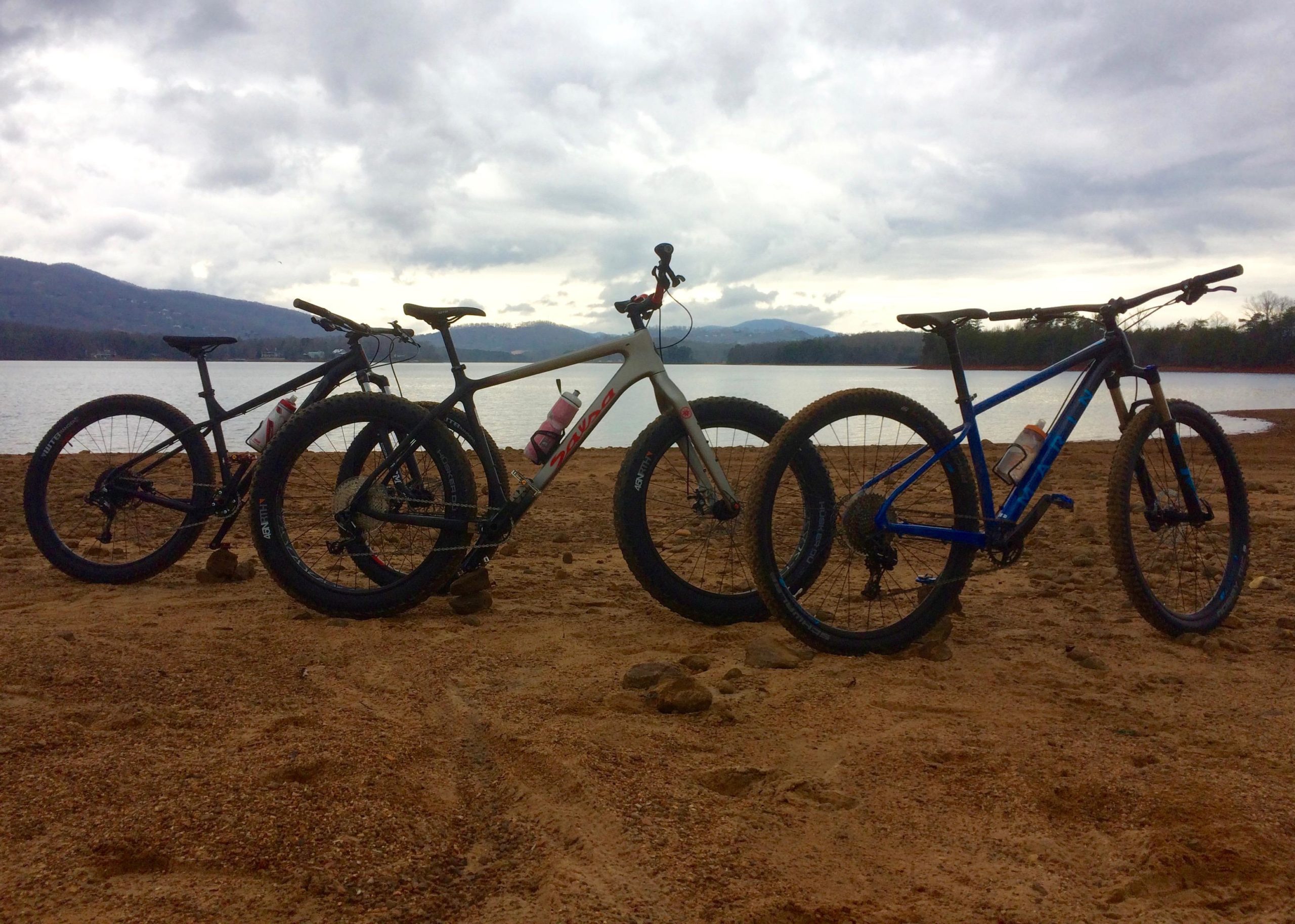 Three mountain bikes are parked on a sandy beach near a lake, with a backdrop of mountains and a cloudy sky. One bike is gray with pink accents, and the other two are black and blue, respectively. In the background, the calm water reflects the overcast sky, creating a serene outdoor scene. Saba Beach mountain bike trail.