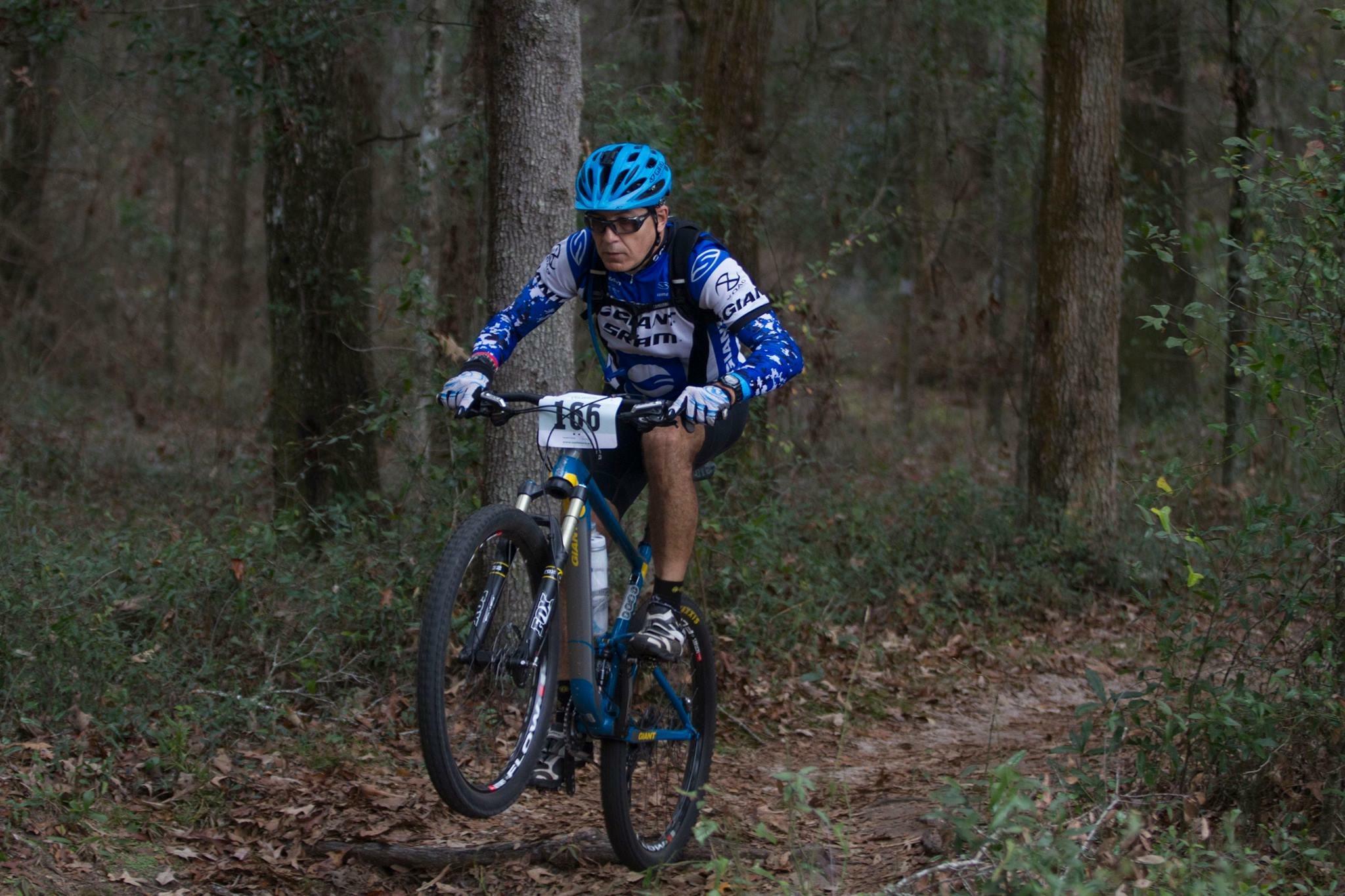 Giant Anthem SX 27.5: A mountain biker performs a jump on a trail surrounded by trees and foliage. The rider is wearing a blue helmet and a long-sleeve jersey, with race number 166 displayed on the front. The bike is a blue mountain bike with thick tires, illustrating an action-packed moment in a natural outdoor setting.