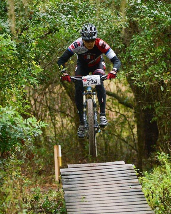 Giant Anthem SX 27.5: A mountain biker in a racing outfit, including a helmet and jersey, performs a jump off a wooden ramp in a forested area. Surrounding greenery enhances the natural setting of the action shot.