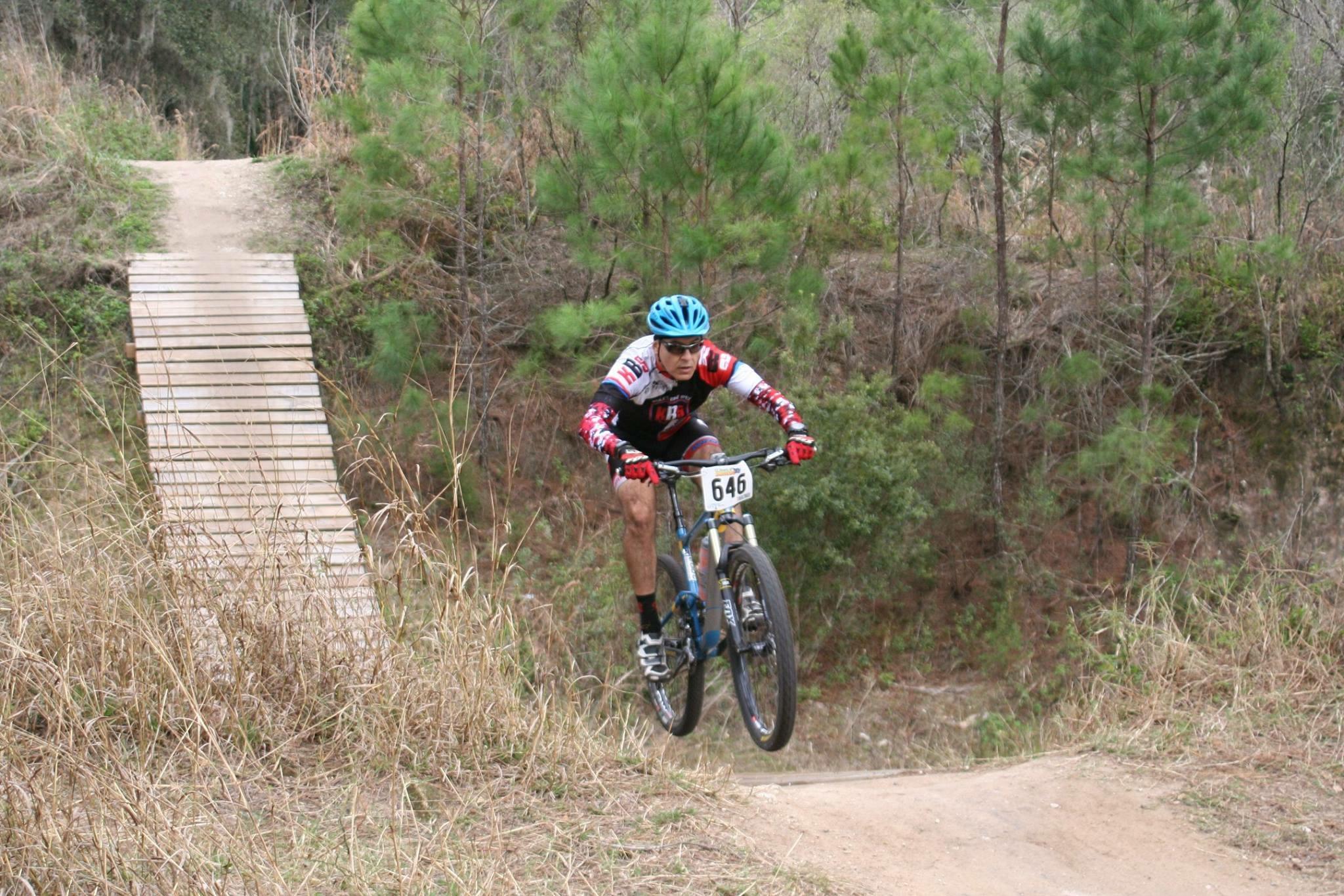 Giant Anthem SX 27.5: A mountain biker performing a jump off a wooden ramp in a wooded area. The cyclist is wearing a blue helmet and a colorful jersey, with a race number displayed. Surrounding terrain includes tall grass and young pine trees.