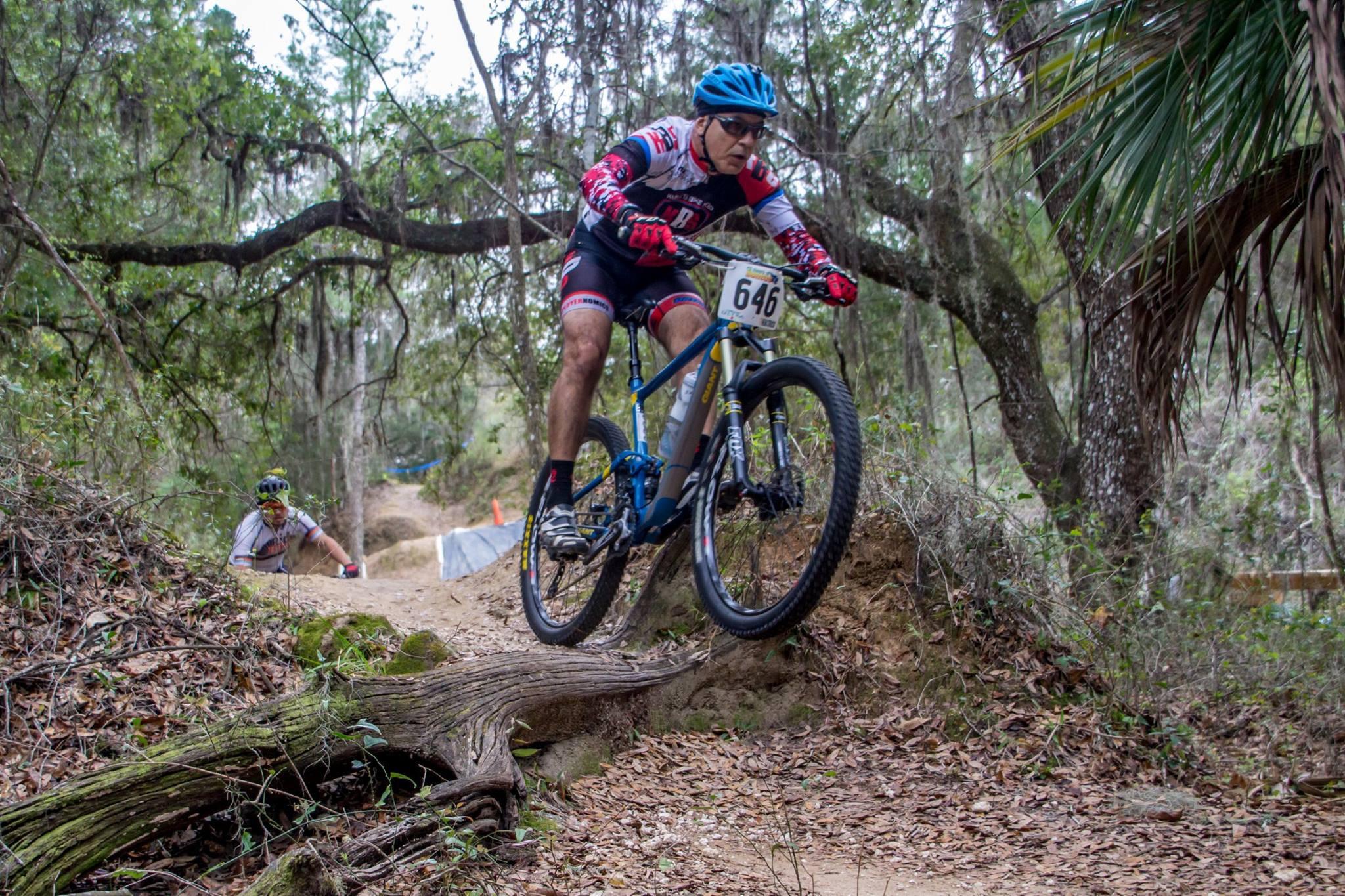 Giant Anthem SX 27.5: A mountain biker in a racing outfit jumps over a log on a dirt trail surrounded by trees and greenery, displaying an action-packed moment. Another cyclist can be seen in the background navigating the trail.