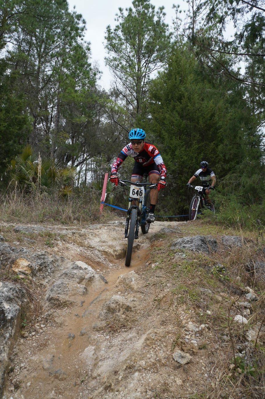 Giant Anthem SX 27.5: Two mountain bikers navigate a rocky trail through a wooded area. The rider in the foreground wears a blue helmet and a brightly colored jersey with the number 646, while the second biker, slightly behind, is dressed in dark gear. The trail is uneven with dirt and rocks, surrounded by grass and trees in the background, creating a rugged outdoor environment.