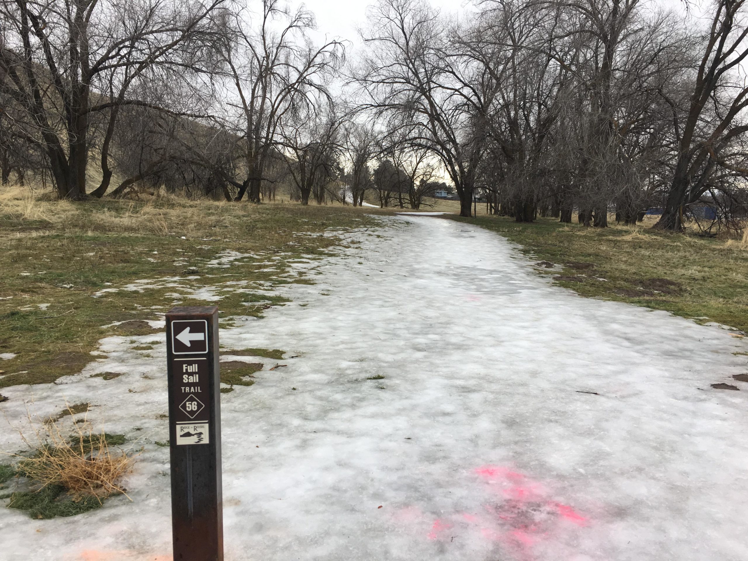 Snow and ice-covered trail sign directing to the Full Sail Trail, surrounded by bare trees and grassy areas. The path appears slippery, with patches of ice and some visible grass. Full Sail mountain bike trail.