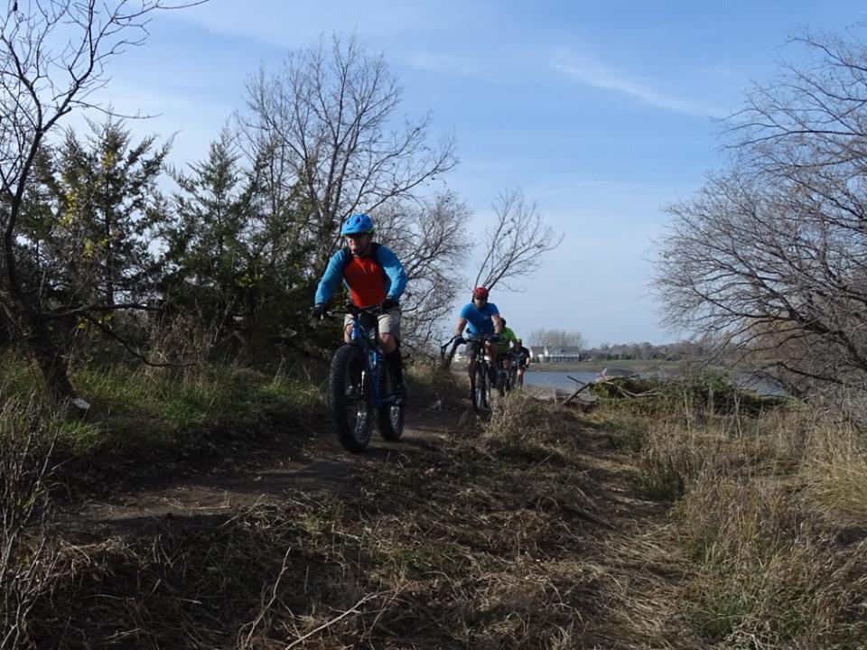 A group of four mountain bikers riding along a dirt trail next to a body of water, surrounded by sparse trees and grass. The riders are wearing colorful cycling gear and helmets, set against a bright blue sky.