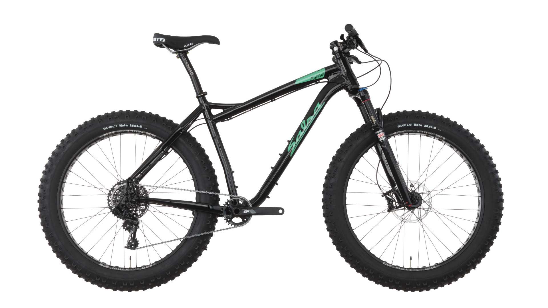 Salsa Mukluk: A black fat tire bike with green and white accents, featuring wide tires and a sturdy frame, displayed against a white background. The bike includes front suspension, a rear derailleur, and a textured seat designed for off-road cycling.