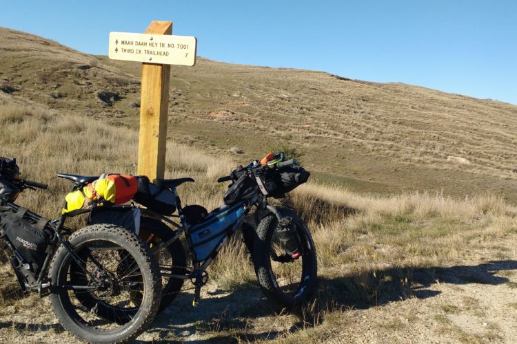Two bicycles with bikepacking gear are parked beside a wooden trail sign labeled “Maah Daah Hey Tr. No. 7001” and “Third Ck. Trailhead” with distance markers. The background features rolling hills and a clear blue sky, indicating a scenic outdoor setting.