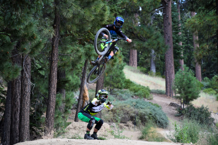 A mountain biker performs a jump in mid-air while another biker crouches on the ground in a forested area. Tall pine trees surround the scene, and the ground is a mix of dirt and sparse vegetation. The flying biker is wearing a blue helmet and vibrant green shorts, while the crouching biker is wearing a black and green outfit with a helmet and goggles.