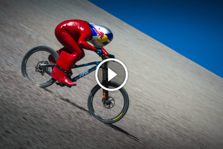 A cyclist dressed in a shiny red suit and helmet is racing downhill on a sleek blue mountain bike. The background features an expansive, slightly angled terrain under a clear blue sky, emphasizing the speed and dynamic motion of the rider.