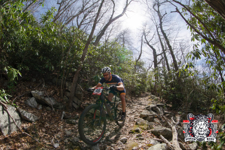 A mountain biker navigates a rocky trail in a wooded area during the Pisgah Stage Race. The cyclist, wearing a helmet and riding gear, displays race number 48 as he skillfully maneuvers over the challenging terrain. The scene is illuminated by sunlight filtering through the trees, highlighting the vibrant foliage around the path.