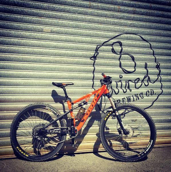 Kona Precept: A vibrant orange mountain bike leaning against a textured metal garage door, featuring a logo for "8 Wired Brewing Co." in the background. The bike is equipped with thick tires and a water bottle holder, showcasing its rugged design suitable for outdoor adventures.